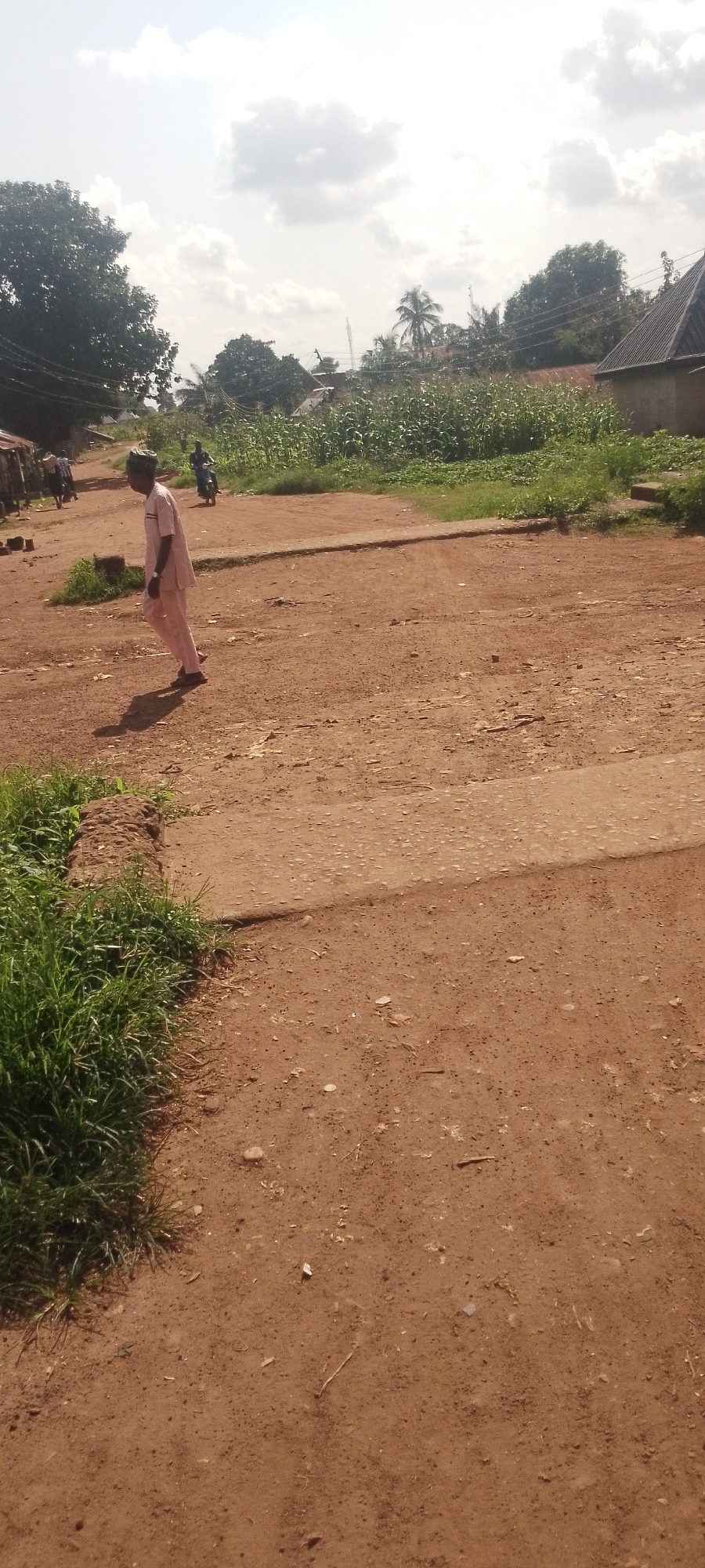 A medium shot shows a dirt road with a man walking on it. He is wearing a pink outfit. The road is dusty and appears to be in a rural or semi-rural area. To the left of the road, there is tall green grass. In the background, there are trees, some houses, and a field of crops, possibly corn. The sky is bright with some clouds. There are other people and a motorcycle in the distance on the road. The overall scene suggests a daytime setting in a warm climate.