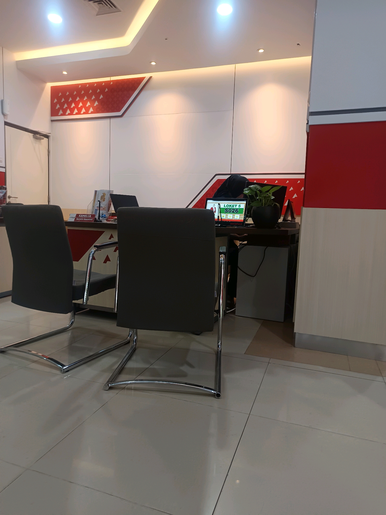 This is a medium shot, eye-level interior photograph taken inside what appears to be a service or administrative office, likely located in Sampang, Indonesia, judging by the text on the screen. The scene is brightly lit by overhead fluorescent and accent lighting.
In the foreground, two dark gray, modern-looking chairs with chrome frames face away from the viewer, positioned towards a counter. The floor is tiled with large, light-colored square tiles, showing some reflection from the lighting.
Behind the chairs is a counter area. On the counter, a laptop is open, displaying a screen with Indonesian text that reads "LOKET 5 S026" and a green graphic. To the right of the laptop, a potted plant with green leaves sits in a dark bowl. A person wearing a dark head covering and black clothing is partially visible behind the counter, interacting with the computer. A smaller black laptop is also visible behind the main laptop.
On the counter, further to the left, a small stand displays printed materials. A name tag or sign on the counter reads "KAMELTA" with the title "Service Assistant" below it.
The background features a wall with a white, smooth finish, broken by a decorative panel on the upper left which has a red and white geometric pattern. The upper part of the wall has integrated white LED lighting. To the right, a vertical section of the wall is red, meeting a white section. A closed white door with a silver handle is visible on the far left of the frame. The overall impression is of a clean, functional, and modern office space.