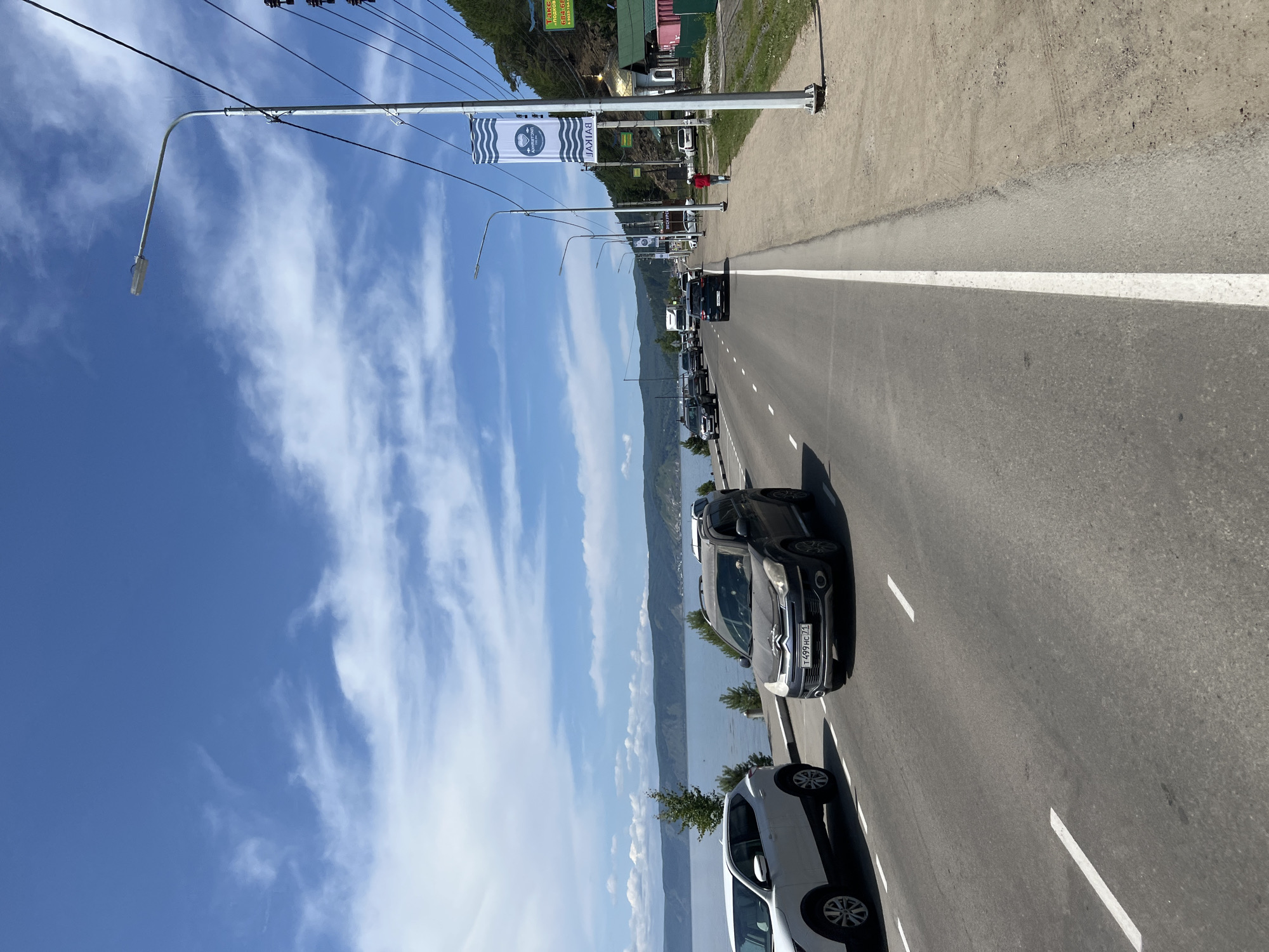A road runs alongside a large body of water, likely Lake Baikal, in Listvyanka, Russia. The sky is blue with scattered white clouds. On the left side of the road, a white SUV is parked. Several other vehicles, including a dark grey SUV with a license plate visible, are driving on the road. On the right side of the road, there is a sidewalk with a lamppost. A banner with wavy lines and the word "БАЙКАЛ" (Baikal) in Cyrillic is attached to the lamppost. Further down the road, there is a small building with a golden dome, possibly a church, and some green and red structures. A person in a red jacket is walking on the sidewalk. The environment appears to be a scenic, coastal road with mountains in the background.