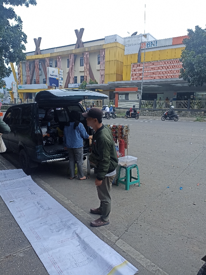 The image depicts a street scene in Jatinangor, Indonesia, located at coordinates -6.933 latitude and 107.773 longitude. A green SUV with its rear door open is parked on the side of the road, facing a large yellow building with a "BNI" sign. A man in a green jacket and khaki pants stands beside the open rear door of the SUV, looking at something inside. A woman in a blue shirt and jeans is standing beside the SUV's rear bumper, seemingly looking at something in the SUV. A long piece of rolled-up blueprint paper is on the ground in front of the SUV and runs parallel to the street. There is a green plastic stool next to a white plastic tub. A red fire extinguisher is leaning against a small, rectangular, cardboard sign. A black motorcycle with two riders is parked behind the SUV, to the right. The street is made of gray concrete and has a light gray painted stripe running down the center.