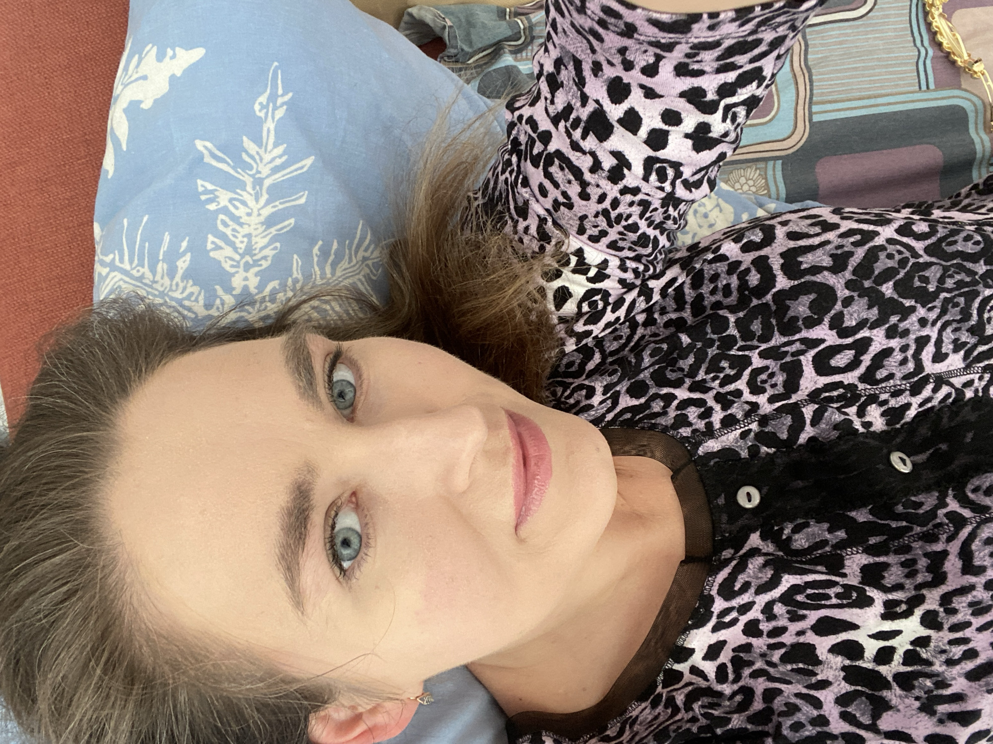 A woman with light skin and blue eyes is lying down. She is wearing a lavender and black leopard print top with a black lace trim and silver buttons. Her hair is brown. She is lying on a blue pillow with a white botanical design. A glimpse of a red surface and a patterned blanket are also visible. It is impossible to determine the location in the city of Yekaterinburg, Russia, or any actions, interactions, or environment from this image alone.