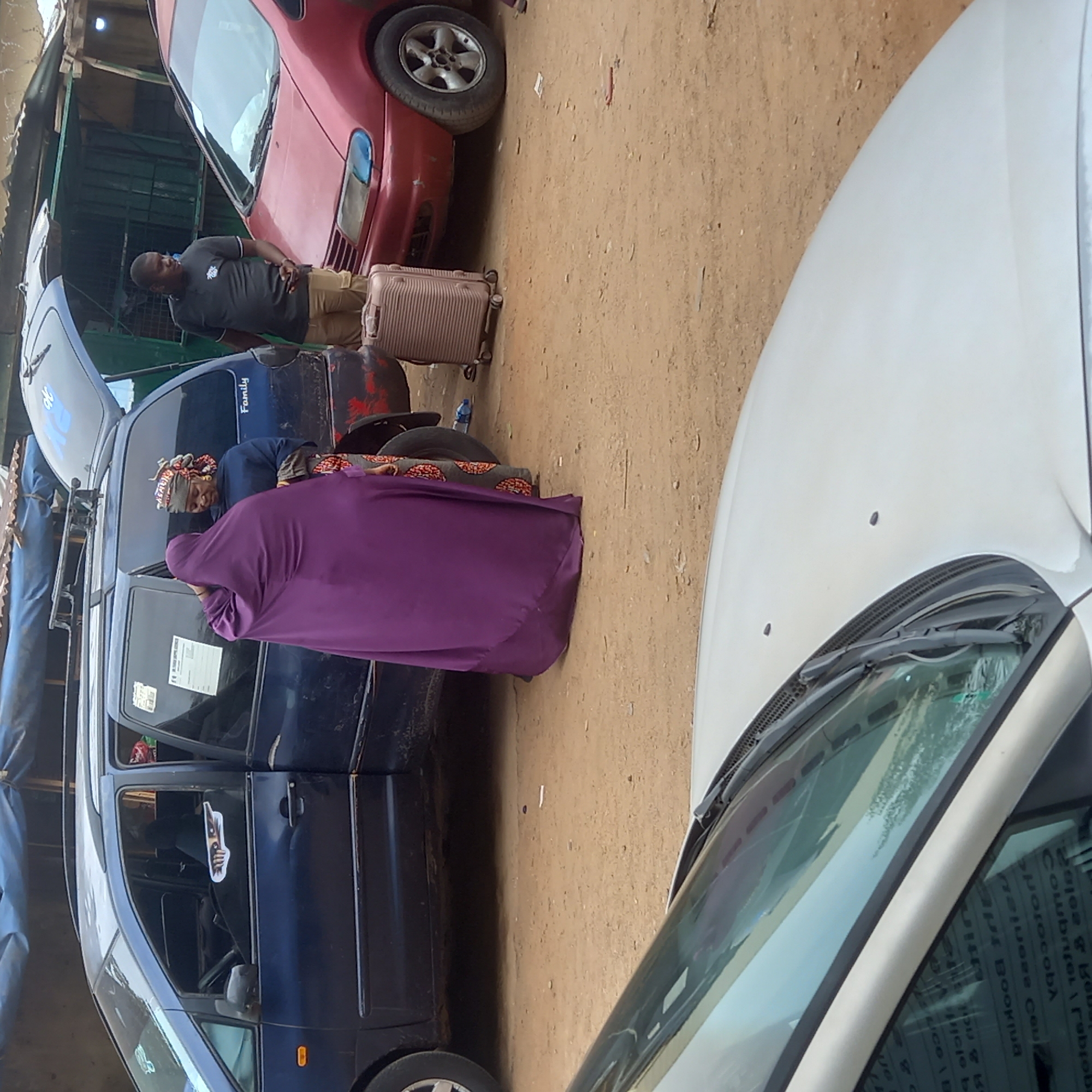 The scene appears to be on a dirt area in what is possibly Dadi Riba, Nigeria. There are three cars visible: a red car, a dark blue car, and a white car. A man in a grey shirt and khaki pants stands near a suitcase and the red car. Two women are near the dark blue car; one is wearing a purple garment. The trunk of the dark blue car is open, and there is a roof rack on top.