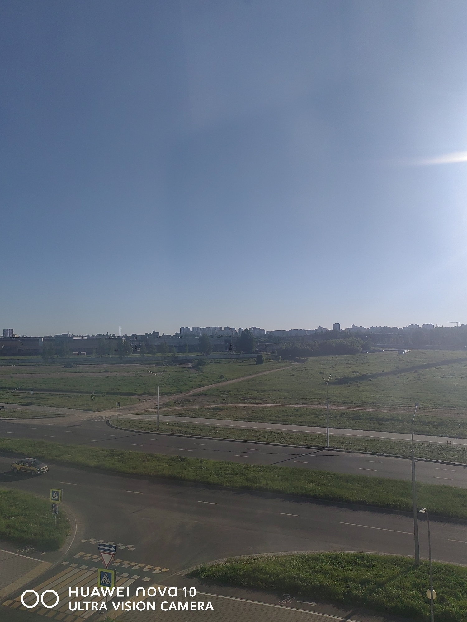The image presents an elevated view of an urban-fringe landscape in Vitsebsk, Belarus, under a clear, bright sky. The scene is dominated by a combination of infrastructure and natural elements.
In the foreground and midground, a network of asphalt roads forms a junction, complete with pedestrian crossings marked by white zebra stripes and a unique checkered pattern, possibly indicating a shared path for cyclists and pedestrians. Traffic signs, including a yield sign, pedestrian crossing signs, and directional arrows, are visible near the crossings. A single yellow car is seen driving on the road to the left. Roadside verges and large, undeveloped green fields with scattered trees extend from the roads towards the horizon. Lampposts are present alongside the roads and within the fields.
Beyond the fields, a distinct city skyline is visible, characterized by a mix of industrial-type buildings and taller residential apartment blocks. The sky above is a uniform, clear blue, with intense sunlight entering from the upper right, creating a bright glare and long shadows across the scene, suggesting either early morning or late afternoon.
No people are explicitly visible, but the presence of pedestrian and possible bicycle infrastructure implies human activity. The overall impression is one of a calm, sunny day in a transitioning urban environment.
A digital watermark in the bottom-left corner reads "HUAWEI nova 10 ULTRA VISION CAMERA," accompanied by a circular logo.