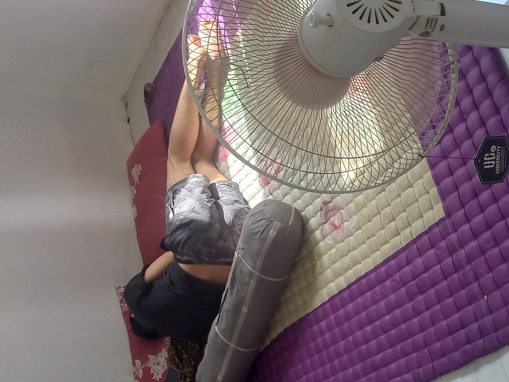 The image captures an overhead, rotated view of a person resting indoors in Puloblang, Indonesia. A white floor fan is prominently positioned directly above the person's upper body and legs, suggesting warm or hot conditions. The person, dressed in a black t-shirt and camouflage-patterned shorts, is lying on their side on a quilted mat.
The mat features distinct purple and cream/beige sections, with the cream part adorned with pink floral patterns. A grey, cylindrical bolster or pillow rests alongside the person. Bright natural light illuminates the scene, casting reflections and shadows, particularly noticeable through the fan's protective cage, indicating it is daytime. The room has plain white walls, suggesting a simple domestic setting. The person appears to be in a state of rest or relaxation. On the purple section of the mat, a black rectangular tag with a white logo and the text "UNDERCITY" is visible.