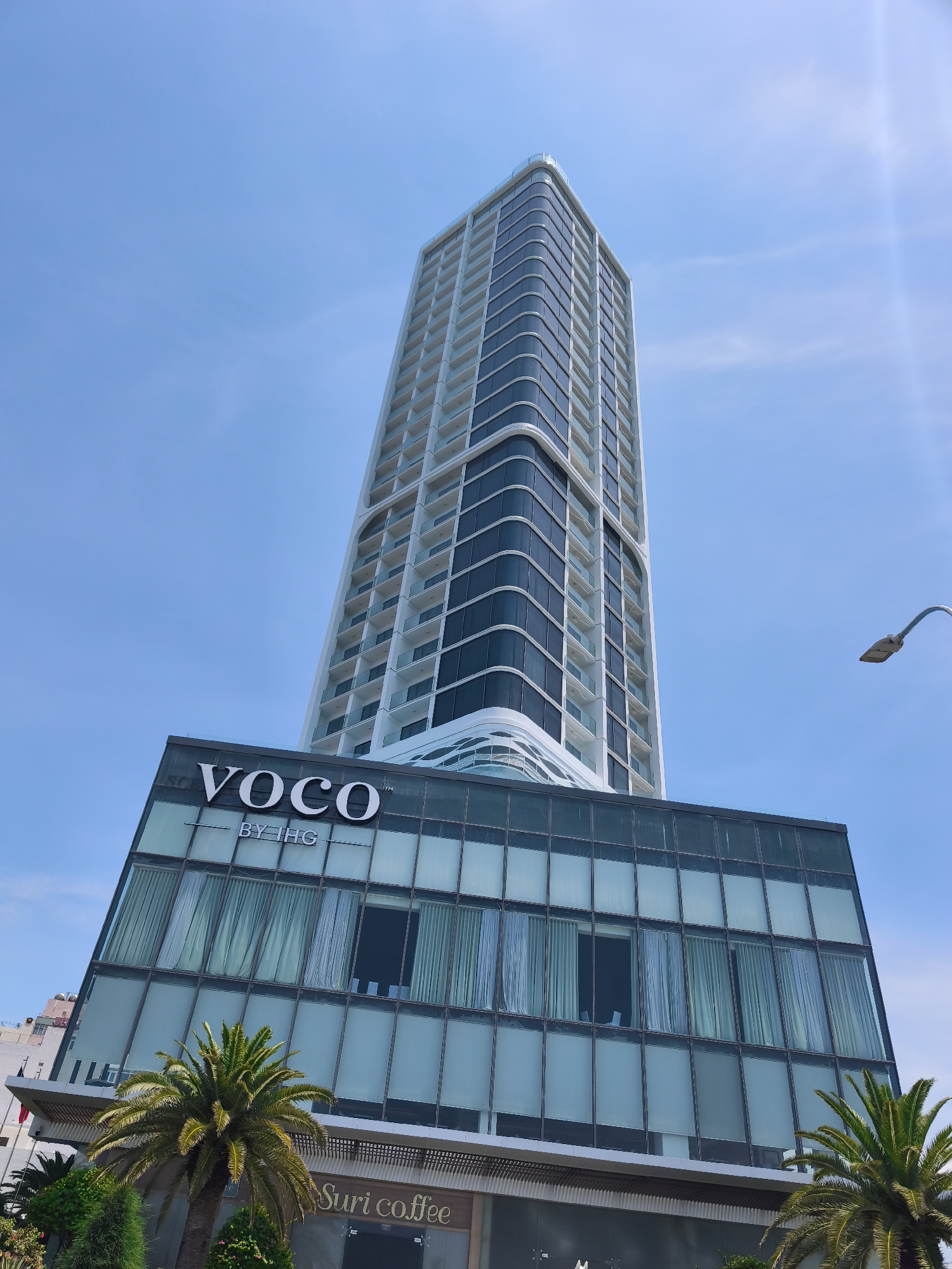 A low-angle, full shot captures a modern, tall skyscraper against a clear blue sky. The building features a sleek, curved facade with alternating bands of white and dark glass. The name "VOCO BY IHG" is prominently displayed in white lettering on a glass section at the base of the skyscraper. Below this, the building's lower levels are clad in dark glass, and at the very bottom, there is a sign for "Suri coffee". Palm trees and greenery are visible in the foreground, suggesting a tropical or subtropical urban environment. A street lamp is positioned to the right of the frame. The image appears to be a photograph of the Voco Nha Trang Hotel, located in Nha Trang Ward, Vietnam.