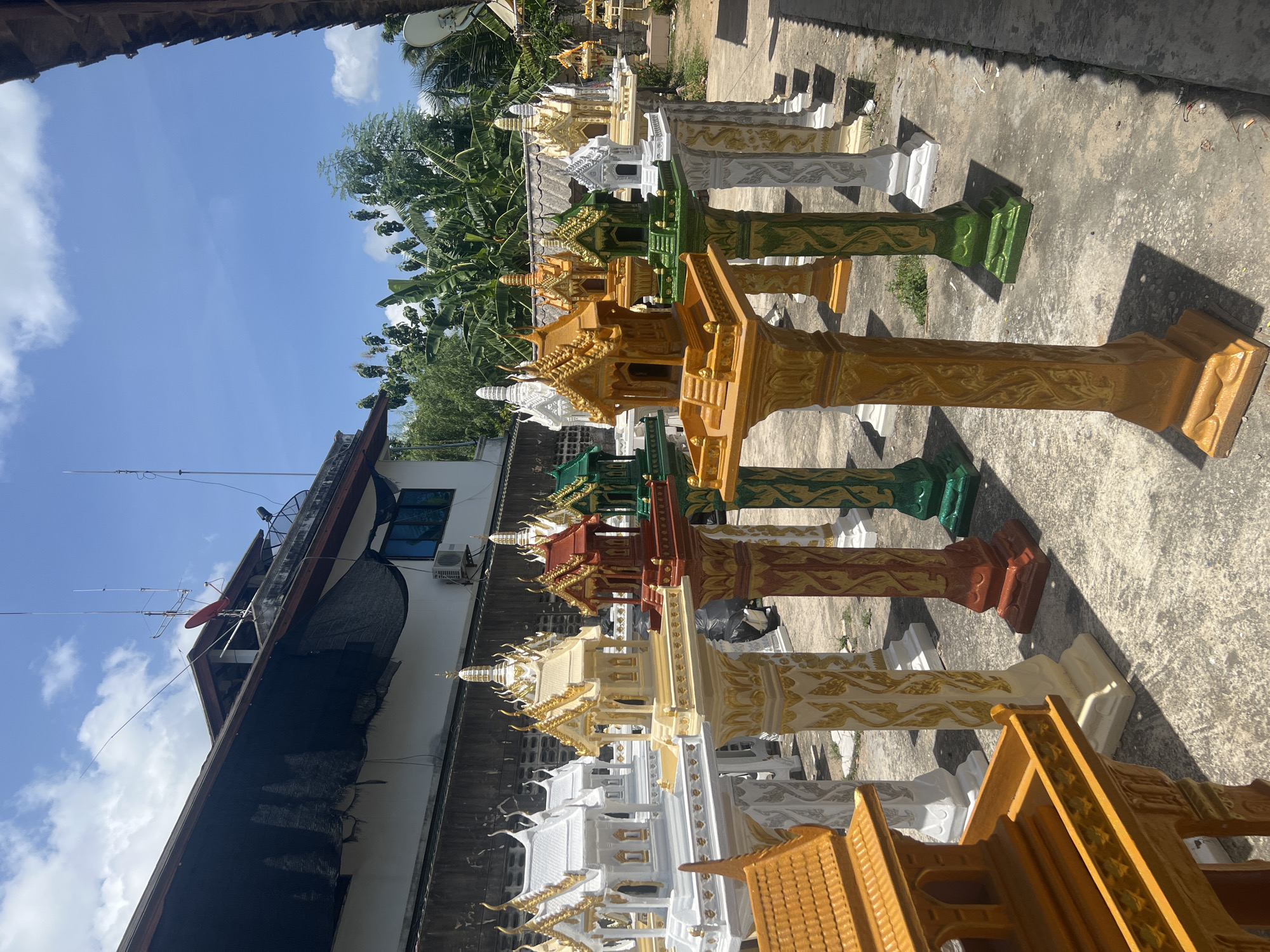 A row of spirit houses, also known as San Phra Phum, are displayed outdoors on a sunny day in Ko Samui District, Thailand. The spirit houses are small, elaborately decorated structures, typically made of concrete or plaster, and painted in various vibrant colors such as white, gold, red, green, and orange. Each house is mounted on a decorative pillar, also painted in coordinating or contrasting colors, and adorned with intricate patterns. The spirit houses are arranged in a line on a concrete ground, with some casting shadows from the bright sunlight. In the background, a building with a dark mesh covering on its roof is visible, along with lush green trees and a clear blue sky with scattered clouds. The scene appears to be an outdoor market or a workshop where these spirit houses are being sold or manufactured. There are no people visible in the image.