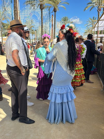 The image captures a vibrant scene in Jerez de la Frontera, Spain. Several individuals are present. Two men stand on the left side of the frame, one in a white shirt, dark pants, brown hat, and sunglasses, and another behind him, wearing a white shirt, a black polka dot tie, a white hat and sunglasses. Three women stand nearby, dressed in traditional flamenco dresses with colorful ruffles. The dress of one woman is light blue with white dots, and she has a white fringed shawl. The other woman wears a purple and turquoise dress. Palm trees and decorative strung lights decorate the background of the scene. The environment appears to be an outdoor fair or festival.