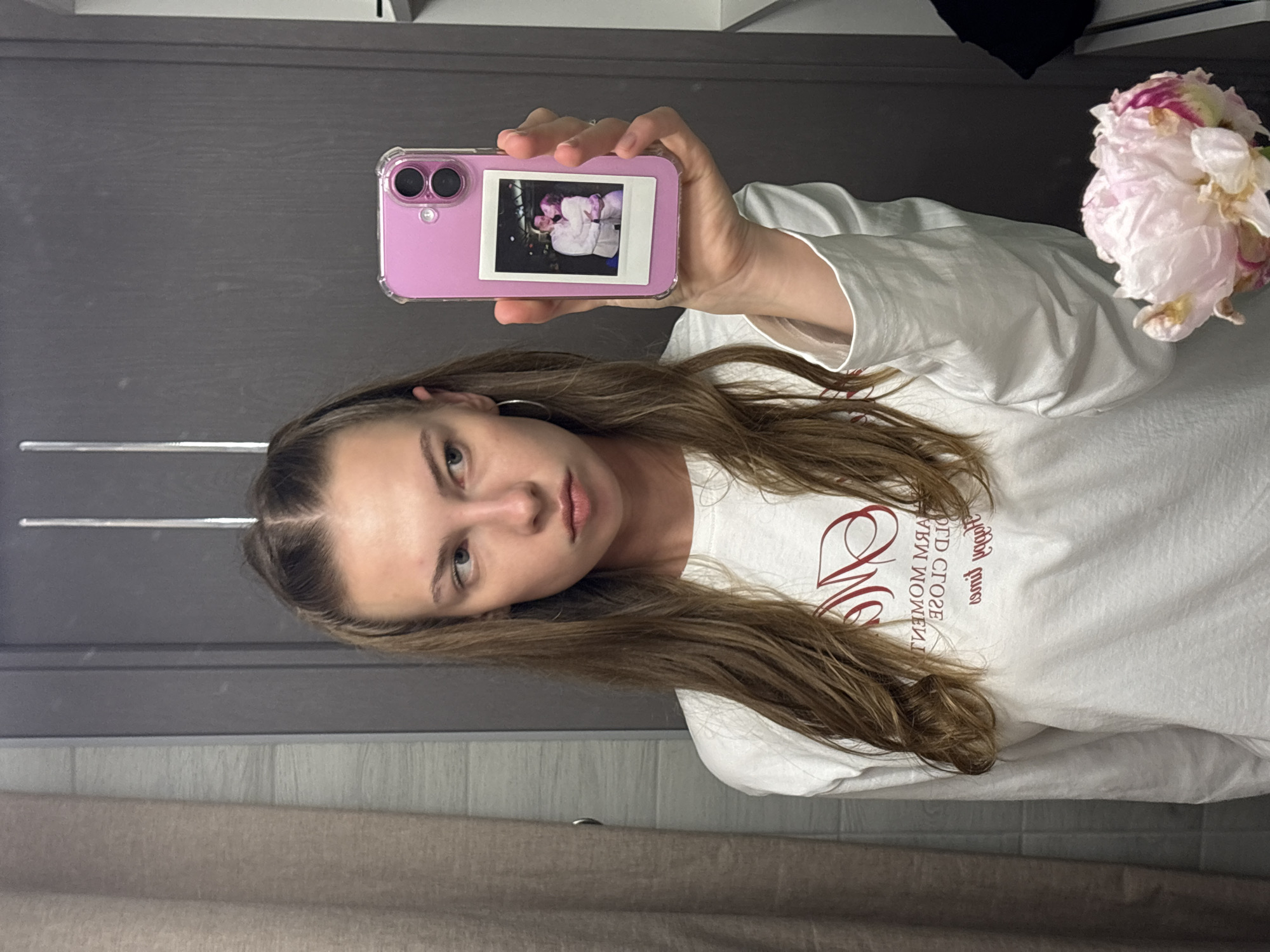 This is a portrait-oriented, slightly blurry, vertical photo taken in Perm, Russia. The main subject is a young woman with long, brown hair, looking towards the camera with a neutral expression. She is wearing a white t-shirt with a red graphic design. She is holding a pink phone case with a polaroid photo inside, showing two people embracing. The background appears to be an indoor setting, possibly a room, with a dark gray door or wall and a light-colored tiled wall. To the right of the woman, there is a partially visible pale pink peony flower. The lighting suggests it is daytime. The text on the t-shirt, when viewed in its correct orientation, reads "LOVE IS THE MOMENT CLOSE".