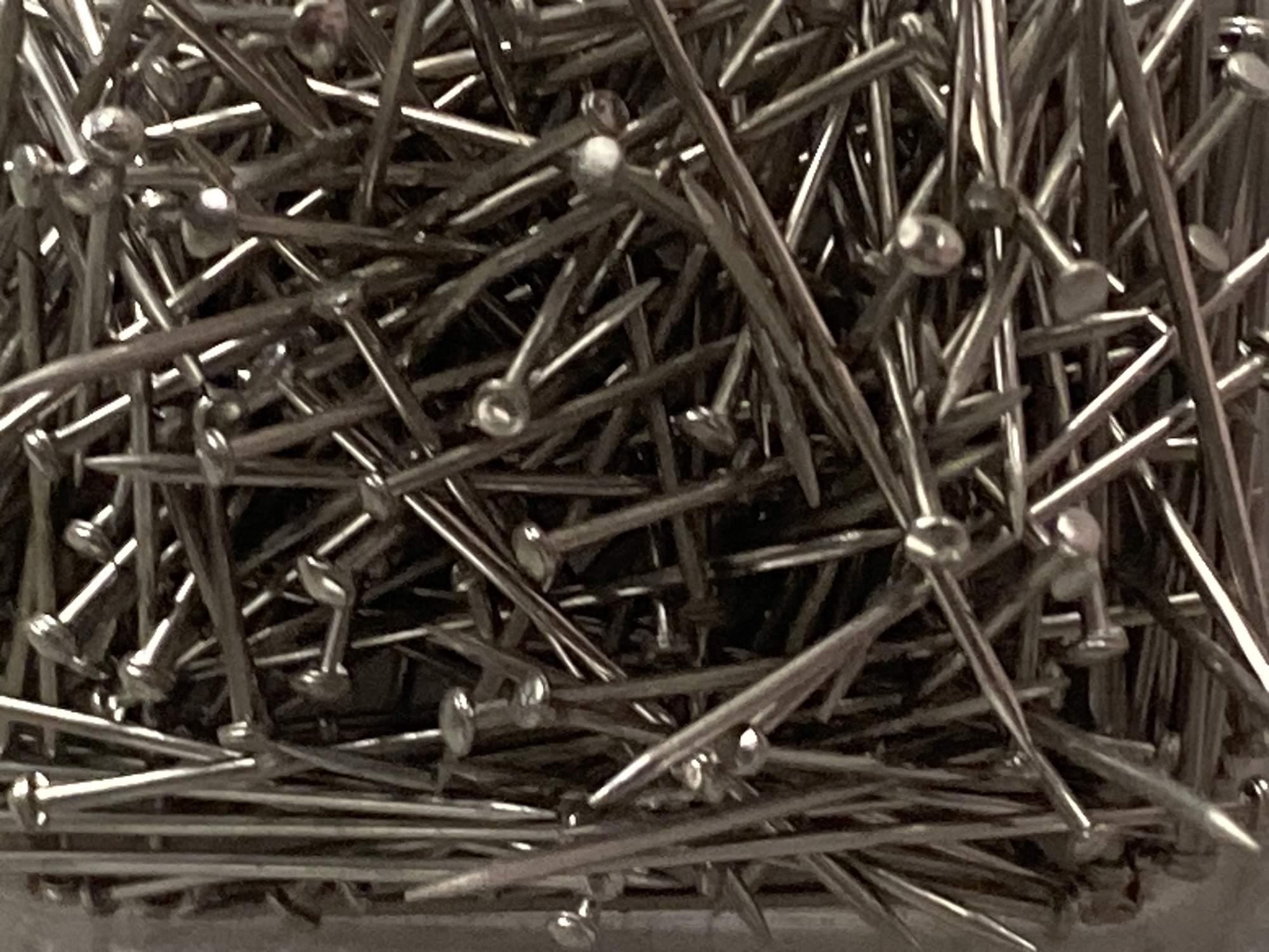 The image is a close-up, overhead shot of a large pile of metal pins. The pins are of various sizes and angles, creating a chaotic yet organized appearance. The metallic sheen of the pins reflects light, adding depth and texture to the image. The background is dark and indistinct, focusing all attention on the pins themselves. There are no people or discernible environmental features visible in the image. The scene is devoid of any contextual information that would allow for its placement in Jauro Yinu, Nigeria, or any other specific location.