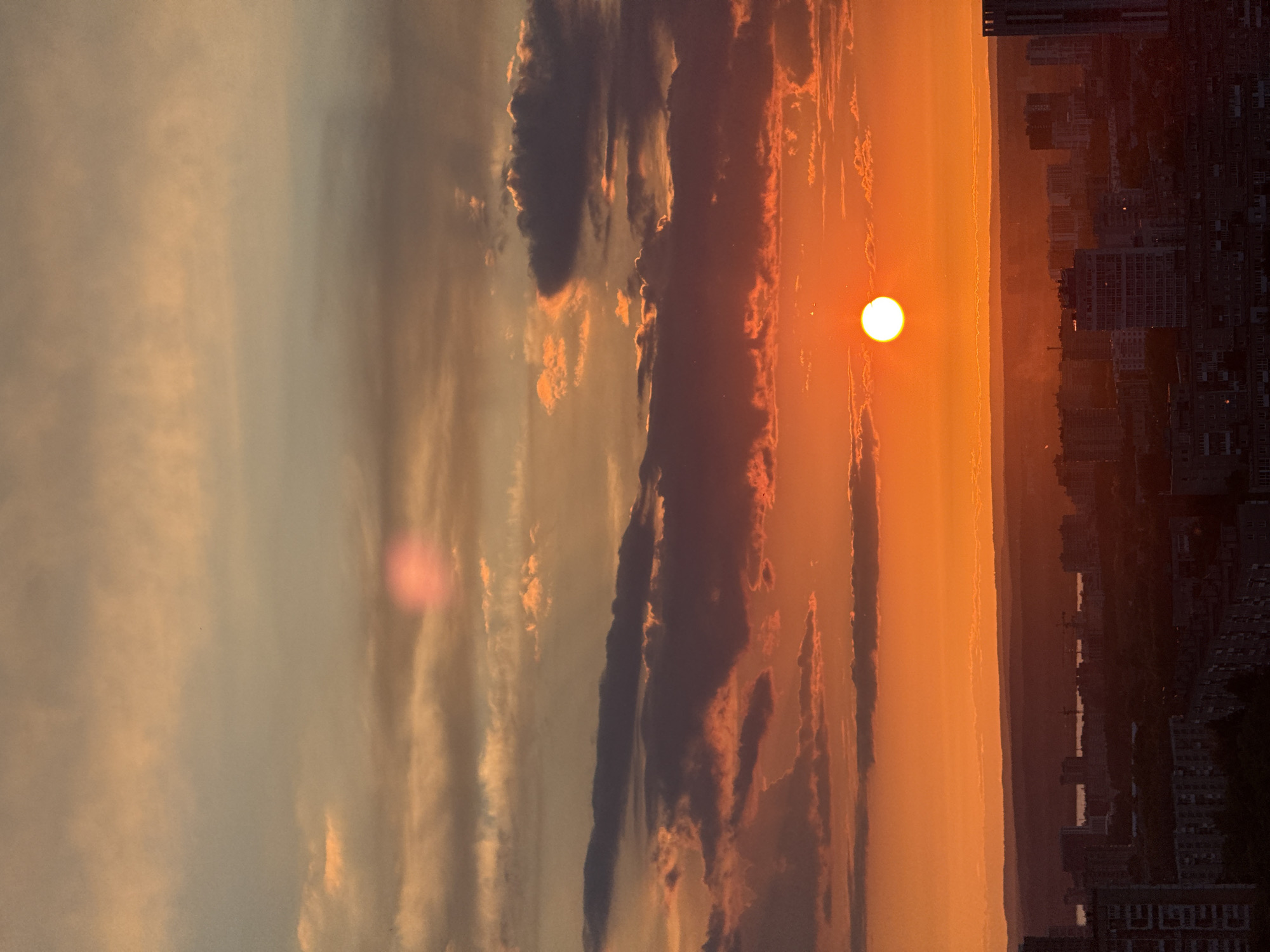 The image displays a vibrant sunset over an urban landscape, likely taken from an elevated vantage point. The bright, circular sun, a stark white orb, is prominently positioned in the right half of the sky, casting intense orange and red hues across the scene.
The sky is a dominant feature, filled with a dramatic interplay of colors ranging from fiery oranges and reds near the sun to softer yellows, greys, and muted blues further away. Various cloud formations are visible; some appear dark and silhouetted against the bright sunset, while others are illuminated from below, catching the warm light. A subtle, pinkish lens flare is visible in the upper left portion of the image.
Below the expansive sky, the dark silhouette of a city skyline stretches across the lower third of the frame. Buildings, mostly indistinct in their shadowed form, create a dense urban tapestry. Beyond the immediate city structures, a wide, dark band suggests a river, valley, or distant plain, with faint glimmering points indicating light reflections or other features. Further in the distance, another strip of land or hills fades into the atmospheric haze.
The scene depicts the end of the day, with the warm, rich colors of sunset dominating the atmosphere. There is no discernible human activity or visible text. Based on the provided context, the location is Сокол, Russia.