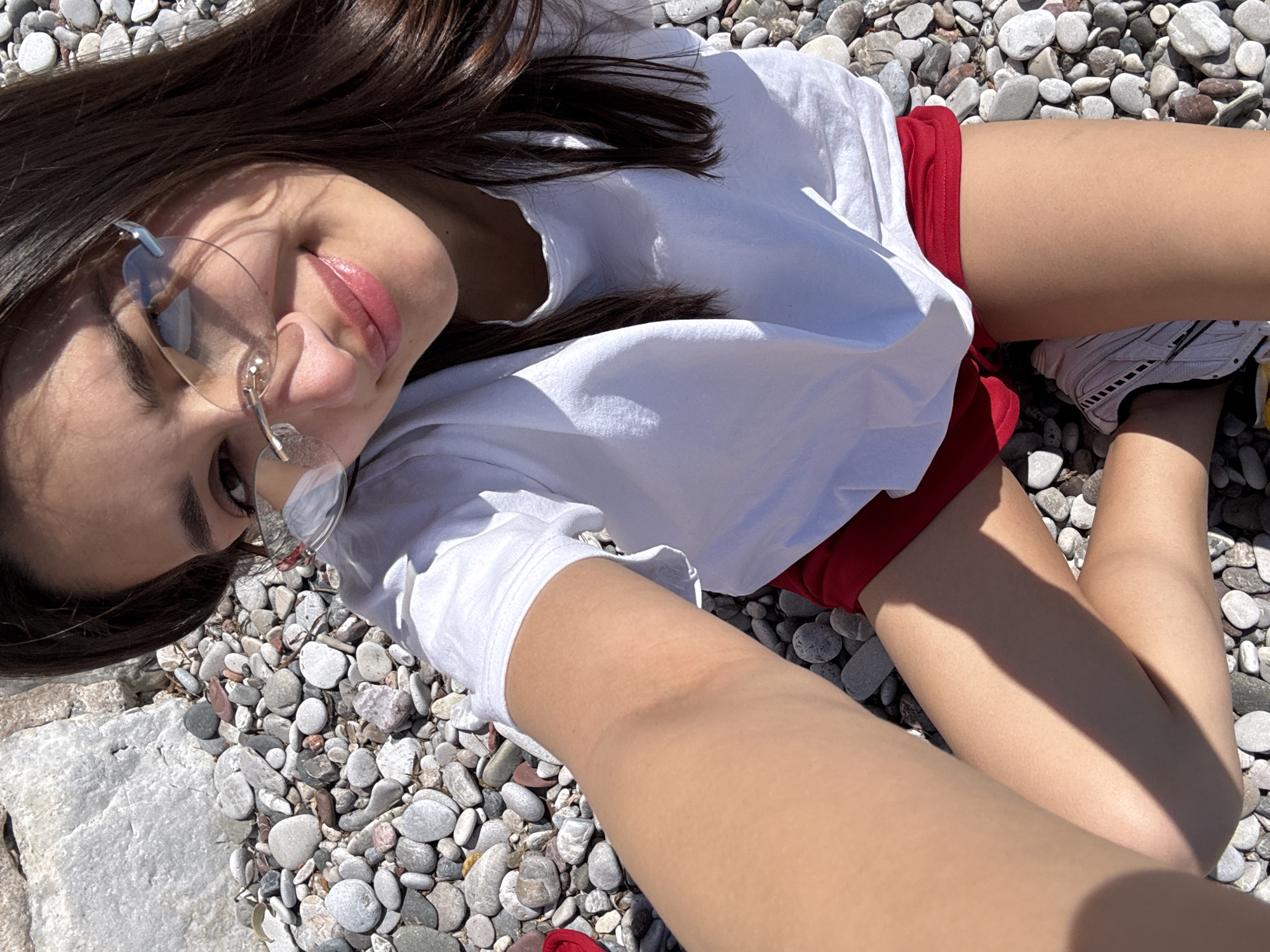 A young woman is lying on a pebble beach in Sutomore, Montenegro. She is wearing a white t-shirt and red shorts. The woman is wearing glasses and is smiling. The background is a pebble beach with a few large rocks. The sky is bright and sunny.