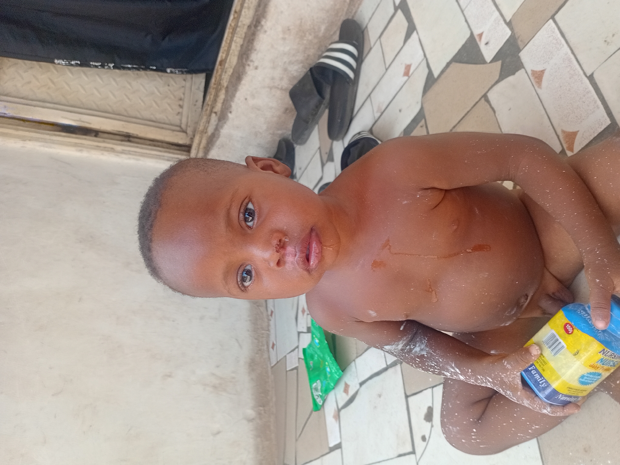 A close-up, high-angle shot captures a young, dark-skinned child seated on a tiled floor, looking directly at the camera with wide, curious eyes. The child's head is shaved, and their body appears wet, with numerous white specks, possibly from water droplets, soap suds, or powder, visible on their skin, arms, and legs. There is also some wetness around the child's mouth.
The child holds a blue and yellow plastic container in their right hand. Visible text on the container includes "NURSE NURSE baby Powder," "100g," and "Family." A small green packet or wrapper is on the floor just behind the child. The floor is covered in irregularly shaped, light-colored tiles with small reddish-brown accents, suggesting a patterned design, and also appears damp.
In the background, two black-and-white striped flip-flops or slippers are visible on the tiled floor. Further back, there's a section of a light-colored wall or structure, and a dark metal grate or patterned surface. A black fabric object is partially visible at the top left of the frame. The overall scene suggests the child is likely bathing or playing with baby powder in a semi-outdoor or wash area, possibly within a home in Lokoja, Nigeria, under bright daytime lighting.