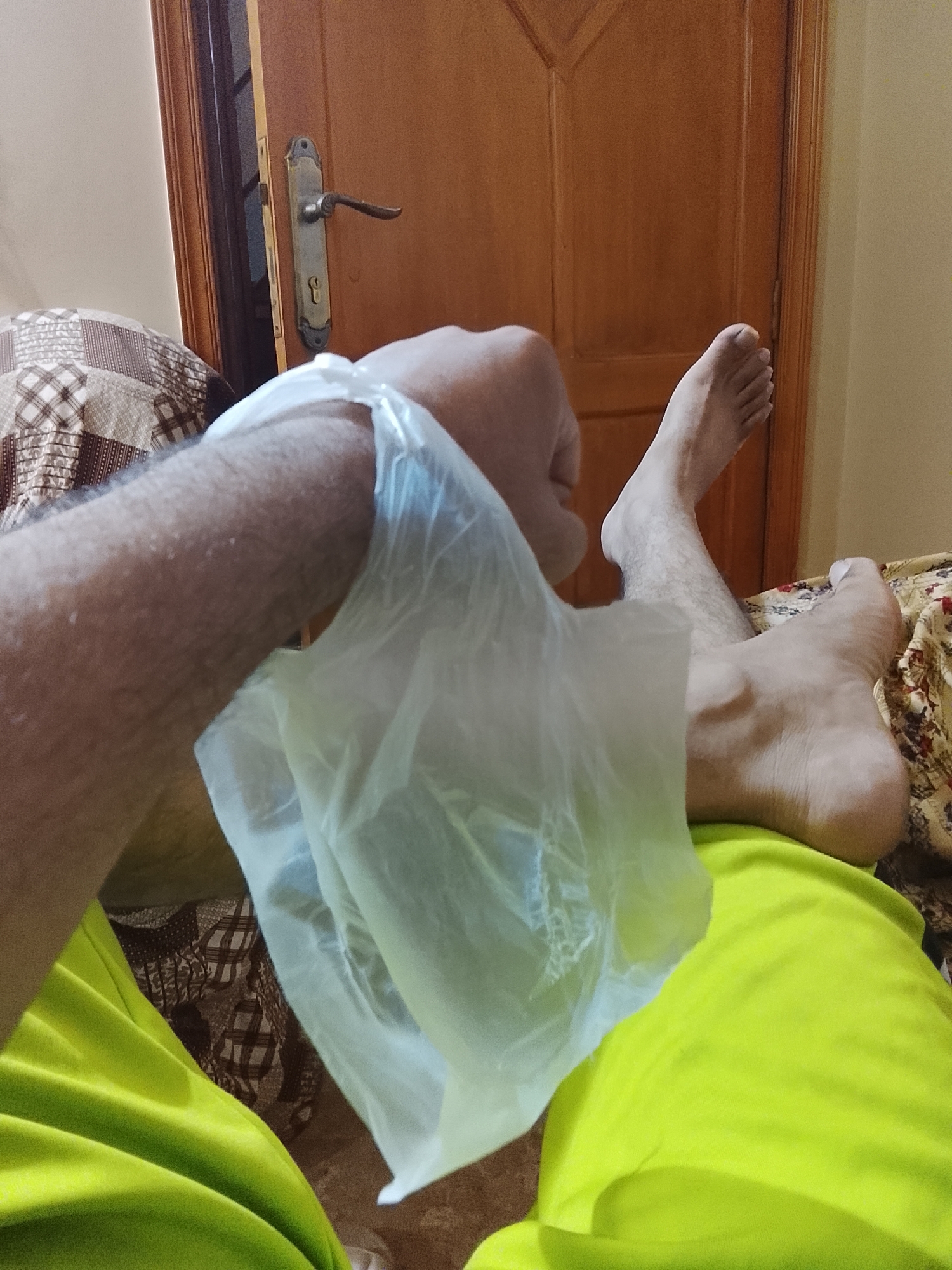 The image captures a first-person perspective of an indoor scene, likely a bedroom or living room. The primary subject is a person, visible through their arm and legs. The arm, covered in hair, is prominently featured in the foreground, holding a translucent plastic bag with the hand inside it. The legs and feet are extended and resting on what appears to be a bed or couch, suggesting a relaxed posture. The person is wearing a bright lime green garment, likely shorts or pajamas.
In the background, a wooden door with an ornate metal handle and keyhole mechanism is partially open, revealing a darker interior or hallway beyond. To the left, a patterned pillow or cushion with a brown and beige checkered design is visible. Another patterned fabric, possibly a bedspread or blanket, is seen under the person's feet on the right side of the frame.
The lighting suggests an indoor setting, possibly with artificial light, creating a soft glow. There are no explicit cues for the time of day or weather. No visible text is present in the image. The activity appears to be a moment of rest or relaxation, with the purpose of the hand being inside the plastic bag remaining ambiguous – it could be for protection, a treatment, or a casual action.