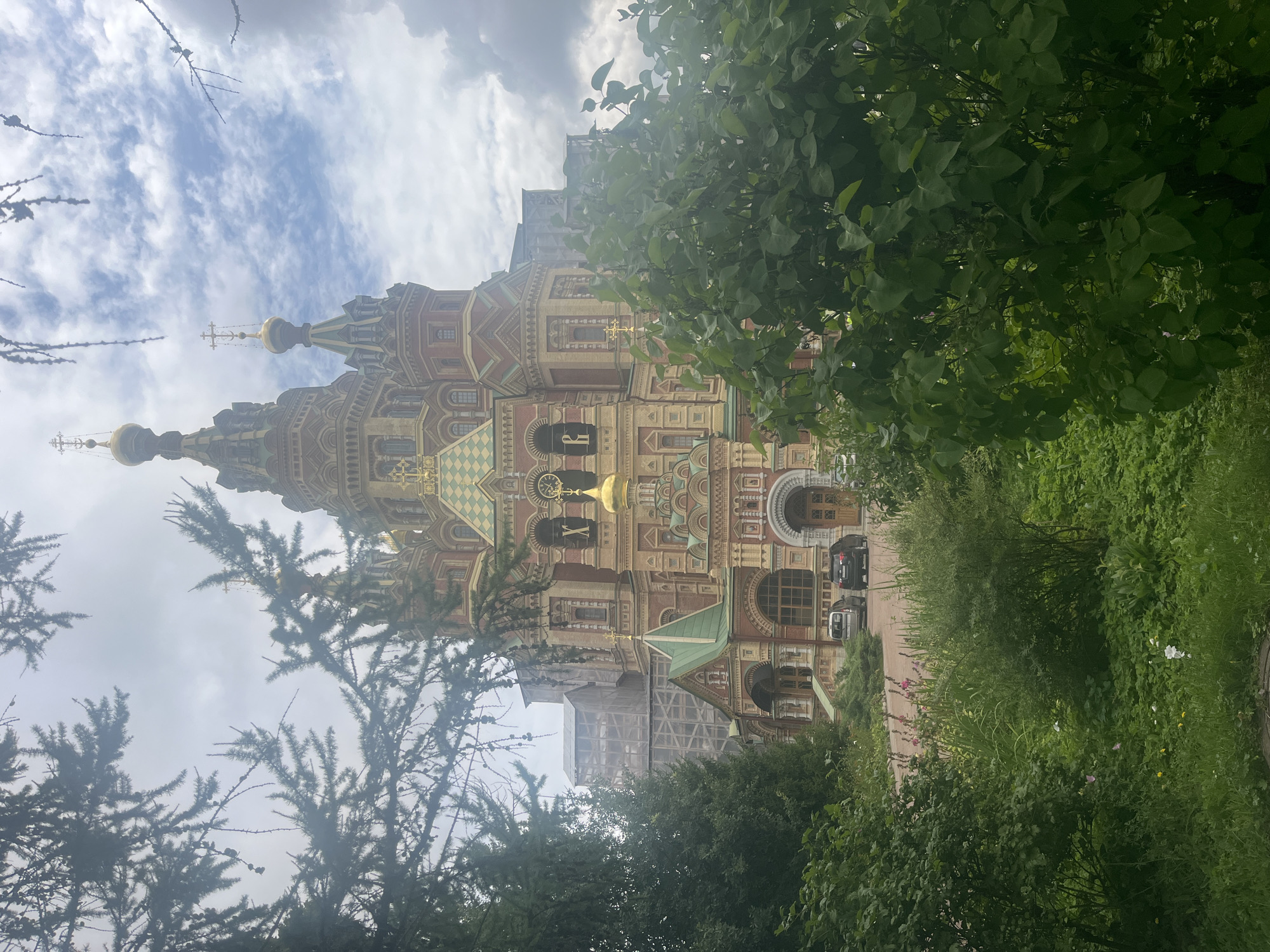 A multi-domed, ornate, reddish-brown and gold Orthodox cathedral is partially obscured by evergreen trees and lush green foliage in the foreground. The cathedral features intricate architectural details, including tiered domes topped with crosses, arched windows, and colorful tile work. Scaffolding is visible on the left side of the building, suggesting ongoing construction or renovation. Two cars are parked on a paved area in front of the cathedral. The sky is overcast with dramatic, dark clouds interspersed with patches of blue sky. The overall impression is one of a grand, historic religious structure set within a verdant, natural landscape under a dynamic sky. The cathedral is the Sobor Petra i Pavla in Peterhof, Russia.