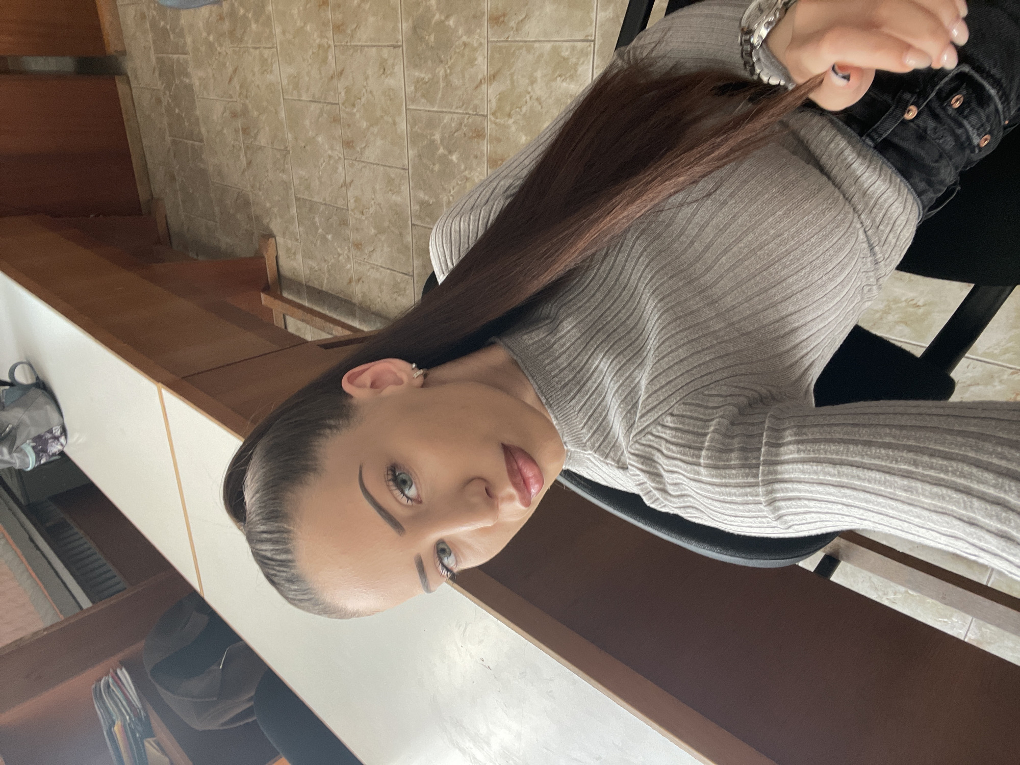 A young woman with long brown hair, fair skin, and light-colored eyes is posing for a selfie. She is wearing a grey turtleneck sweater and black pants with visible copper-colored rivets. She is sitting in a black chair and appears to be indoors. The background includes a brown wooden surface, possibly a desk or shelving unit, with a white surface above it. A wall with light brown and beige tiles is also visible. There is no indication of the location in Veliko Tarnovo, Bulgaria, within the image.