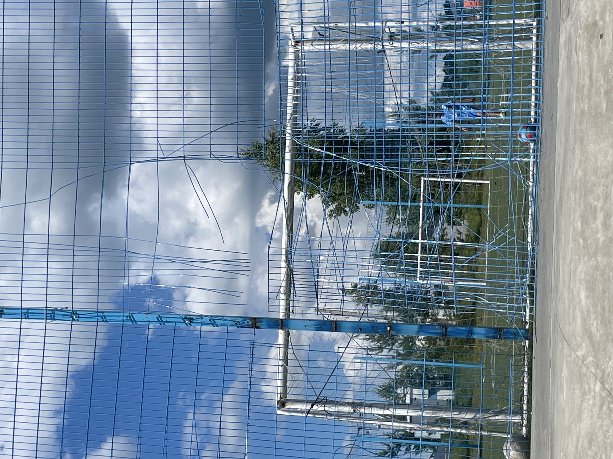 An outdoor sports field, possibly a football pitch, is visible in Laishevo, Russia, under a cloudy blue sky. A prominent blue metal fence, partially damaged with wires hanging loose, occupies the foreground, obscuring parts of the scene. Through the fence, two football goals with white posts and netting are visible. In the middle ground, a young person wearing a blue and white patterned shirt and blue shorts stands near one of the goals. A football lies on the field in front of this individual. Another football rests on the ground to the left of the frame. Trees and buildings are visible in the background.