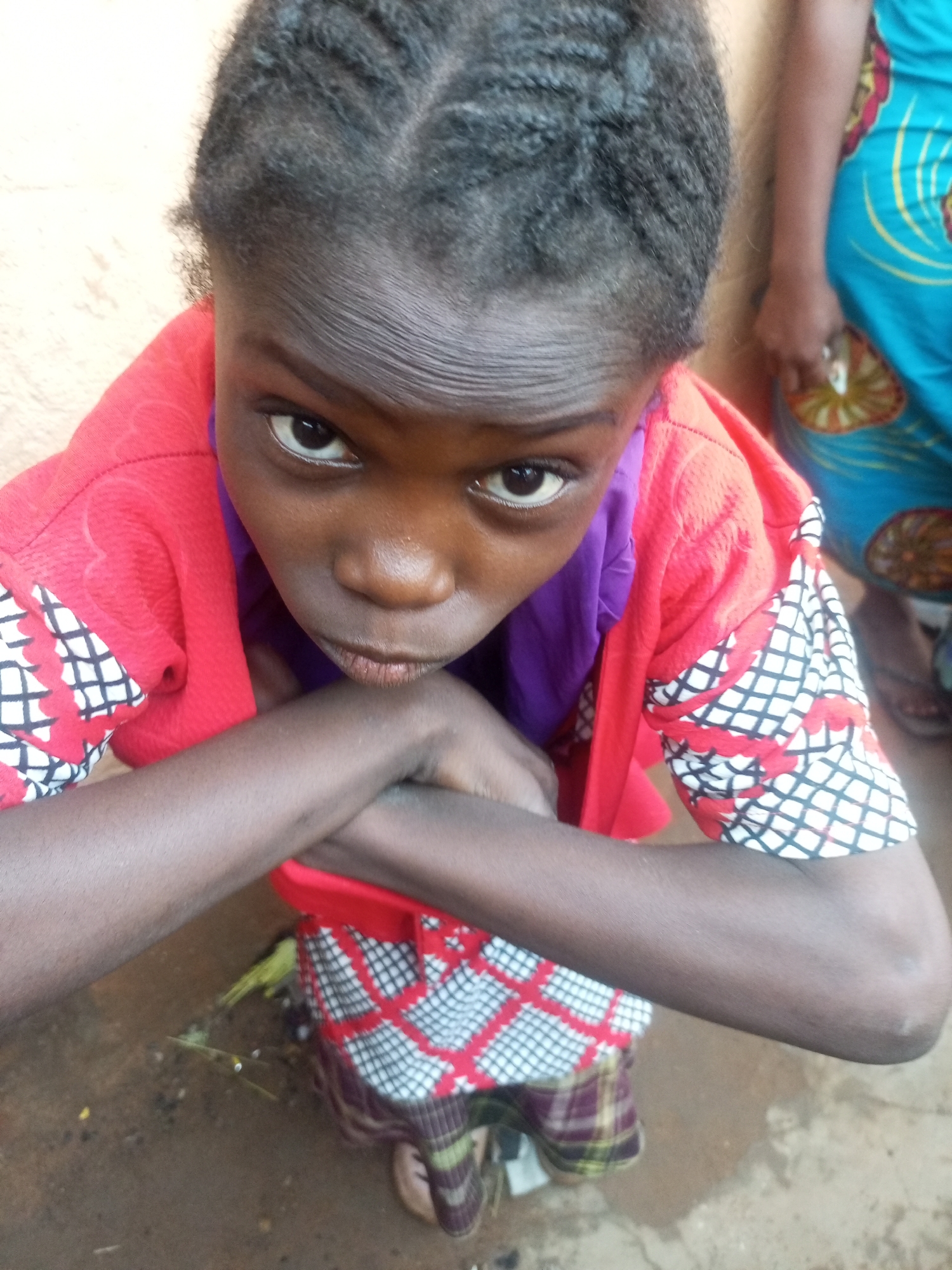 A high-angle, close-up shot shows a young Black person, likely a child, with braided dark hair and dark skin. They are wearing a red top with white and black patterned sleeves and a purple garment underneath. Their arms are crossed in front of their chest, and they are looking directly at the camera with a serious expression. The background is blurred, but appears to be an outdoor setting with a rough, dirt ground and a textured, light-colored wall. Another person, out of focus, is visible to the right, wearing a blue and patterned garment. The scene is likely located in Tempure, Nigeria.