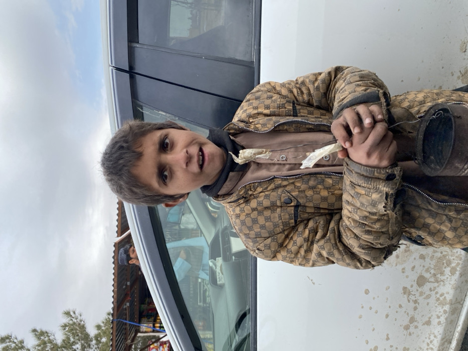 A young boy, approximately 8-12 years old, with short brown hair and an open-mouthed expression, stands in the foreground, looking directly at the viewer. He wears a soiled, brown and tan patterned quilted jacket with visible tears on the cuffs, over a light brown long-sleeved shirt and a black neck gaiter. His hands hold a small, dark metal can or bucket with a wire handle, and several small, light-colored items, possibly candies or seeds, strung together in plastic wrappers.
Behind the boy, a white car with dark tinted windows is positioned; its lower body panel is splattered with dirt. In the background to the left, a roadside stall with a corrugated metal roof is visible, displaying various items. A male individual, wearing a light blue traditional Afghan shirt and a dark cap, is partially visible in reflection within the car's window and behind the car's A-pillar, seemingly attending the stall. A section of a coniferous tree is also visible on the far left. The sky is overcast with scattered clouds, indicating an outdoor, possibly cool, environment. The scene is located in the city of Kandahar, Afghanistan, near a roadside market or thoroughfare.