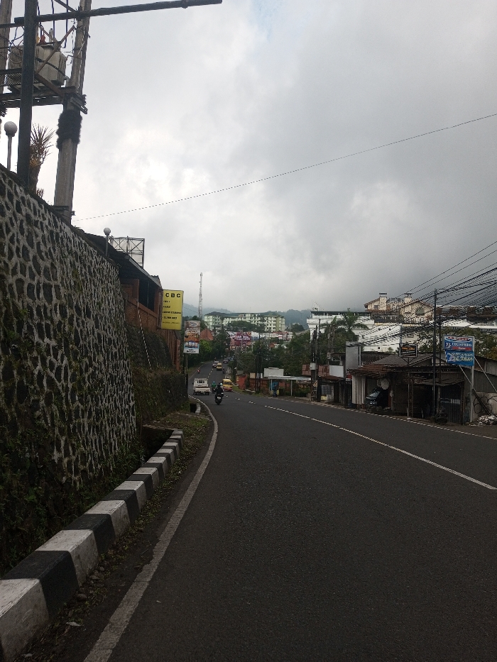 A road curves through a hillside town under a cloudy sky. A tall stone wall runs alongside the road, marked with a black and white curb. Buildings line the opposite side of the road, interspersed with power lines and signs. In the distance, taller structures can be seen nestled in the hills. Traffic moves along the road, including cars and motorbikes. A utility pole stands prominently on the left side of the frame.