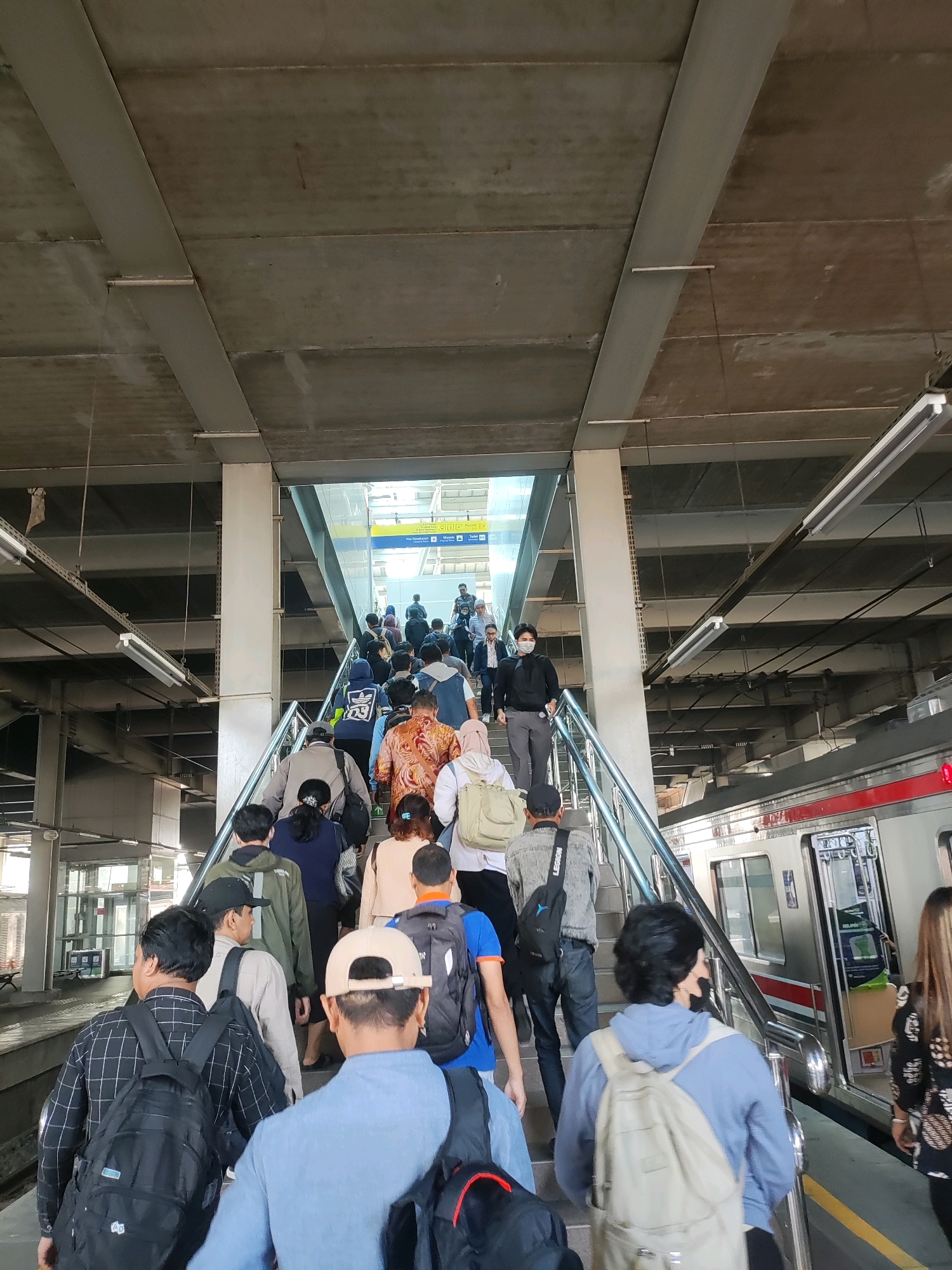 The image depicts a busy scene at a train station, likely in Bekasi, Indonesia, as indicated by a sign in the background with text possibly in Indonesian. A crowd of people are ascending a wide set of stairs leading to an elevated platform. The architecture suggests an indoor-outdoor environment with a concrete ceiling supported by large beams and pillars. Fluorescent lighting fixtures are visible along the ceiling. The people are diverse in age and attire, many carrying backpacks, suggesting they are commuters or travelers. Some individuals are wearing face masks, a common practice for public health. At the bottom of the stairs, a train is visible at a platform to the right, with its doors open. The train appears to be silver with red accents. The overall atmosphere is one of movement and daily transit.