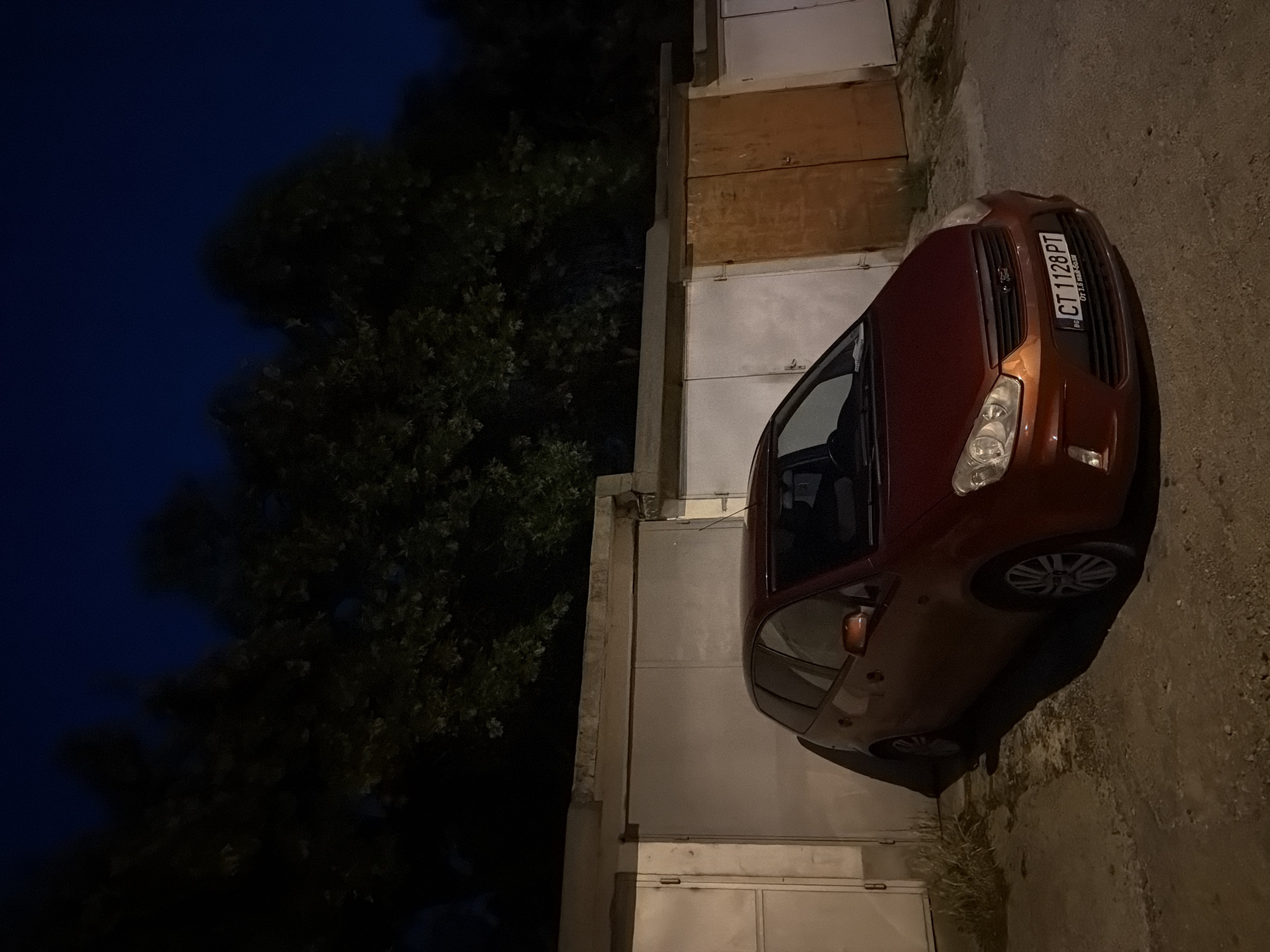 At night, an image captures a reddish-brown metallic Ford C-Max parked facing slightly right in front of a row of garages in Stara Zagora, Bulgaria. The car's front license plate, "CT 1128 PT," is clearly visible, indicating Bulgarian registration, with "BG" on the left and a sticker below reading "От 1.6 най-добър." The vehicle is illuminated by an unseen artificial light source, casting shadows on the ground.
Behind the car, a series of individual garages constructed of concrete are visible. From left to right, the first two garage doors are light-colored, possibly painted metal. The third garage door is distinctively brown or orange, appearing to be made of wood. A fourth, lighter-colored garage door is partially visible on the far right. The concrete structure above the garage doors shows signs of age, with visible weathering and some cracks.
The ground in the foreground is an uneven surface of asphalt or concrete, with scattered dirt and sparse patches of dry grass near the garage walls on the left. In the background, dark silhouettes of dense trees rise against a deep blue night sky. No individuals or discernible activities are present in the frame.