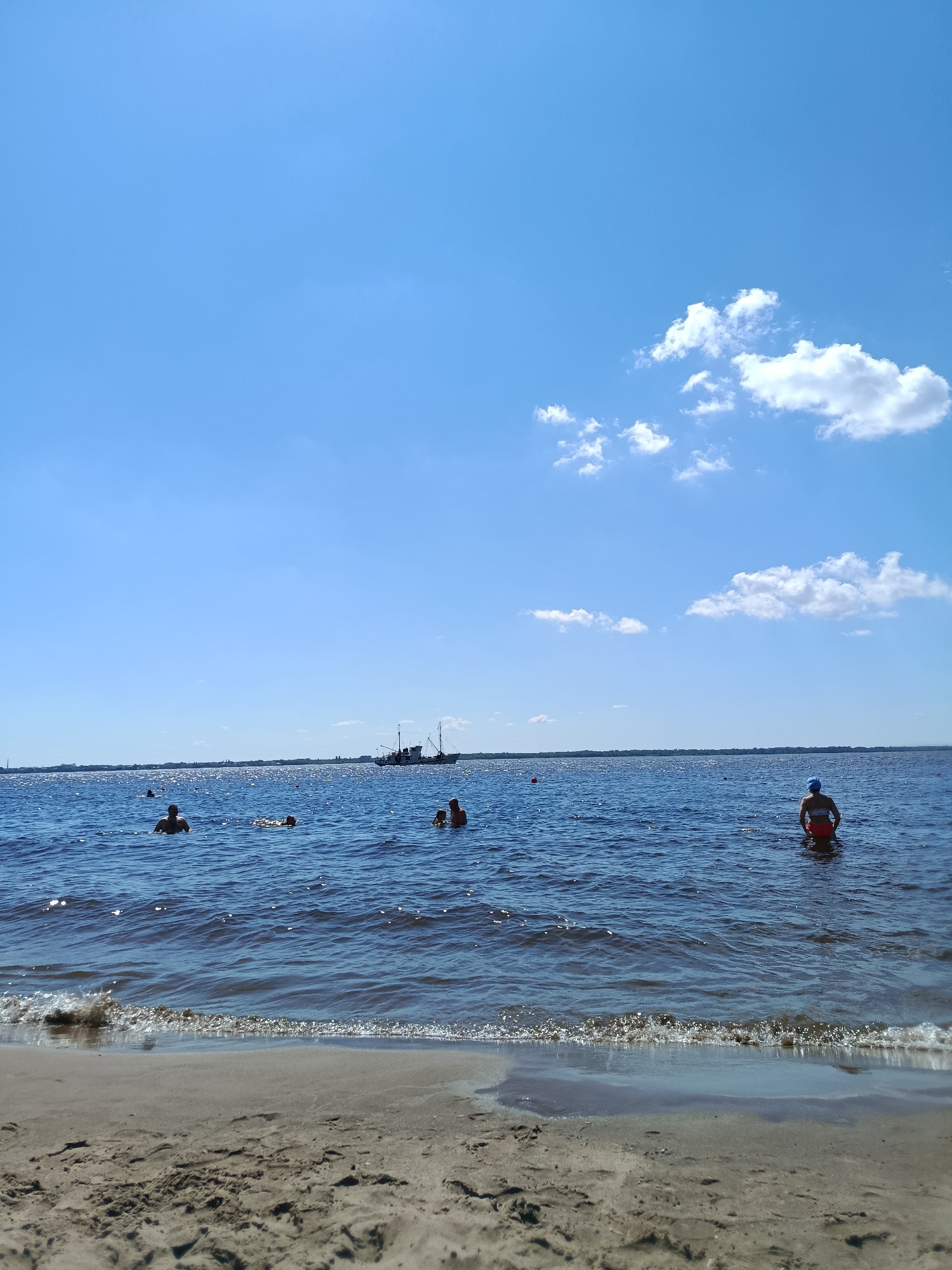 The image captures a sunny day at a beach, likely in Saratov, Russia, given the presence of the Volga River which borders the city. The foreground shows a sandy shore with gentle waves lapping at the edge. Beyond the water, several people are visible swimming or wading. In the middle ground, a large ship is anchored on the horizon, with a hazy skyline of buildings suggesting an urban area. The sky is a clear blue with scattered white clouds. The overall scene depicts a typical summer day at a waterfront recreational area.