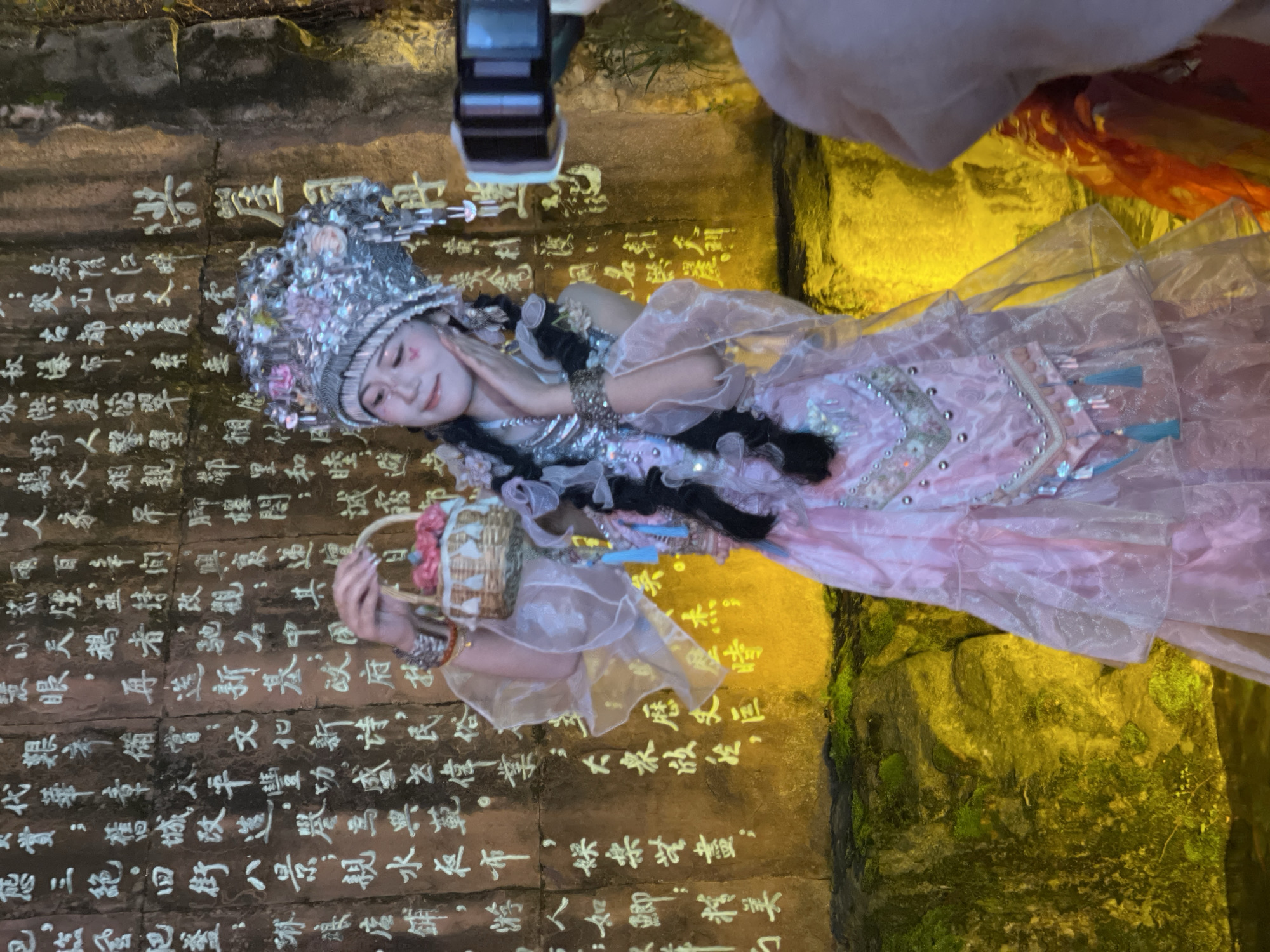 The image shows a young female in an elaborate, traditional Chinese costume posing against a rock wall in Yuzhong District, China, likely at a culturally significant site featuring carved rock formations. The female subject, of East Asian descent, wears a complex silver-colored headdress adorned with pink floral motifs, glitter, and dangling ornaments. Her attire includes a multi-layered gown in light pink and translucent white fabric, featuring intricate silver embroidery, beadwork, and light blue tassels. Her face is made up with blush and small red heart-shaped markings below her eyes. She holds a small woven basket containing pink artificial flowers in her raised left hand, while her right hand is positioned gently near her jawline. Two prominent dark braids are visible under her headdress.
To the right of the primary subject, a second individual, partially visible, holds a black smartphone or small camera horizontally, aiming it towards the costumed female, indicating a photoshoot or video recording.
The background is a textured rock wall, extensively covered with white Chinese characters that appear carved or painted. The lower section of the wall exhibits green moss. The scene is illuminated by strong yellow-orange artificial light originating from below, casting a warm glow on the rock wall and the lower parts of the subject's costume. A dark, reflective surface is visible at the bottom left, consistent with water or a wet surface.