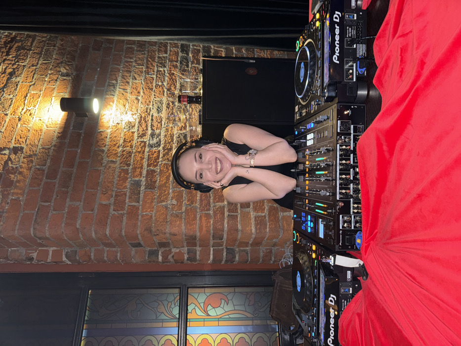 The image depicts an indoor scene in an entertainment venue in Moscow, Russia, featuring a smiling woman positioned behind a professional DJ setup.
The primary subject is a female in her late 20s to early 30s, with dark hair, wearing a dark sleeveless top or dress. She has black over-ear headphones on and is smiling broadly at the viewer, with her hands cupped under her chin, resting on the DJ equipment. A silver-toned watch is visible on her left wrist, and a small tattoo is on her left forearm.
The DJ setup consists of two Pioneer DJ CDJ-3000 media players flanking a central Pioneer DJM-900NXS2 mixer. The equipment is illuminated by various operational lights. The foreground is dominated by a swath of vibrant red fabric, draped over a surface, partially obscuring the lower portions of the DJ console.
The background features an exposed red brick wall, lending an industrial aesthetic to the space. Mounted on this wall is a black, cylindrical up-down wall light fixture casting warm illumination. To the right of the woman, a dark cabinet or shelf holds two clear glasses with ice and a small red bottle, possibly a soft drink. To the left, a multi-paneled window with stained glass-like decorative inserts displays intricate patterns in yellow, orange, blue, and green hues. A brown leather or suede bag rests on a surface near this window. Dark curtains are visible on the far right edge of the frame. The overall environment suggests a bar, club, or lounge setting.
