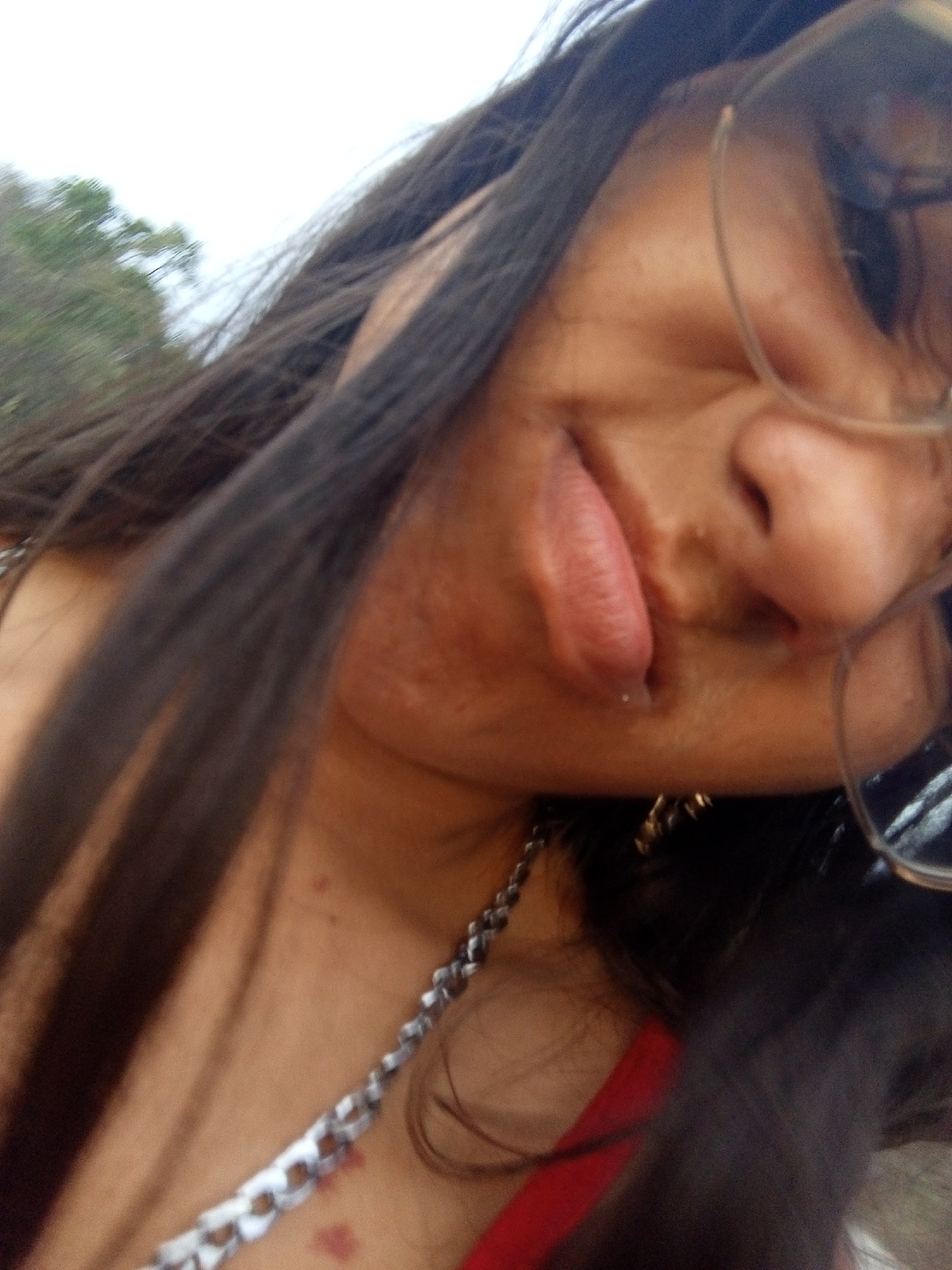 The image presents a close-up, angled portrait of an individual with medium-brown skin and long, dark hair. The subject's head is tilted counter-clockwise, with the right side of the face and left shoulder prominent. The individual wears eyeglasses with thin, light-colored frames, partially obscuring the right eye. A silver-colored link chain necklace is visible on the left side of the neck and chest. A section of a red garment, possibly a top or strap, is visible on the lower left. The person is actively sticking their tongue out, which is elongated and protrudes from the mouth. A small, light-colored piercing or stud is present on the lower lip, slightly to the left of the center. The skin exhibits some texture irregularities on the cheek and jawline, and faint red marks are visible on the upper chest/collarbone area. The background is out of focus, showing a bright, light-colored sky in the upper left and blurred green foliage consistent with an outdoor environment. The specific location within the city of Juan Griego, Venezuela, cannot be determined from the visual information presented in the image.
