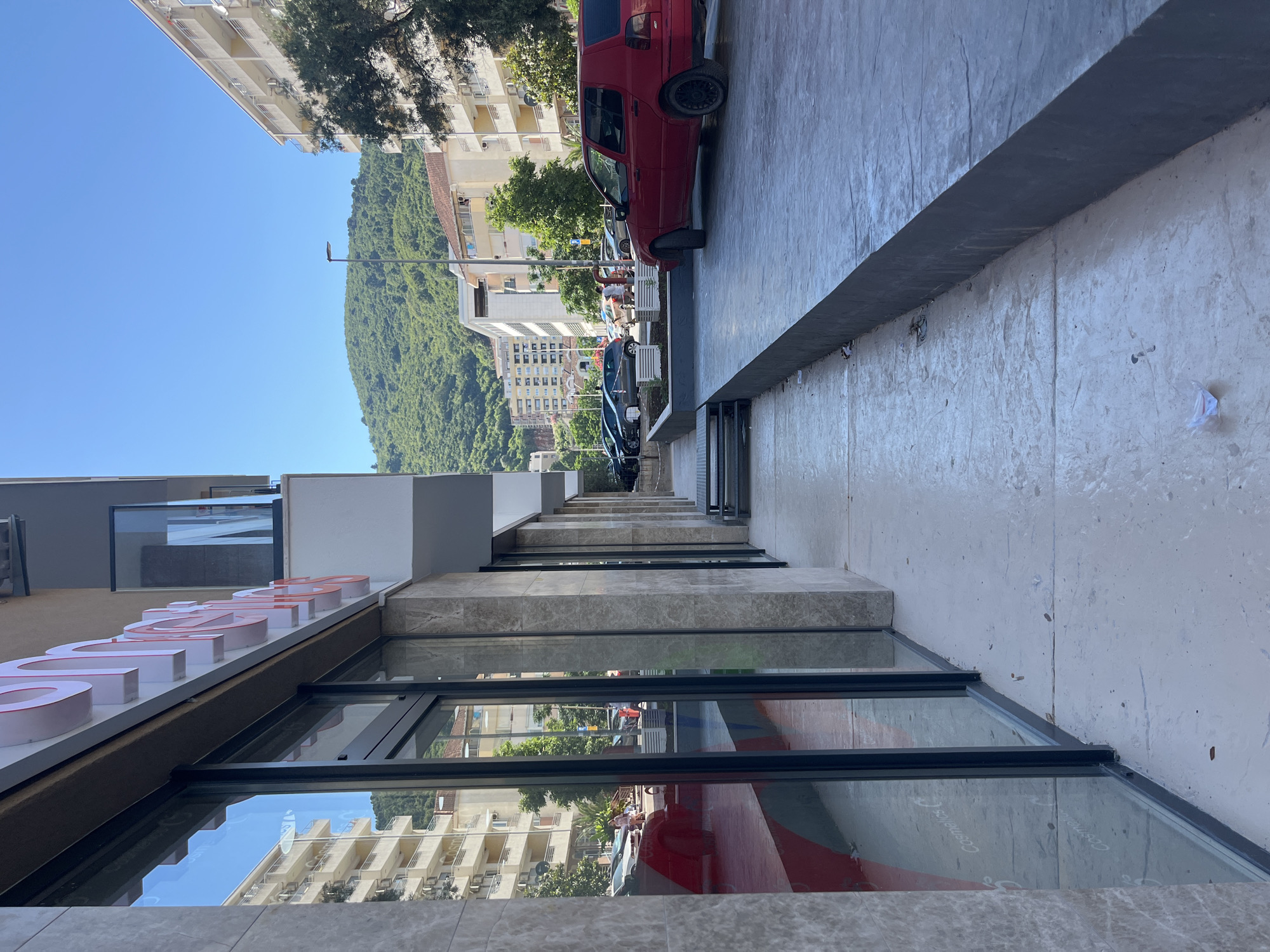 The image depicts a street-level view of a commercial building in Budva, Montenegro. The building features large glass windows, some of which reflect a multi-story residential building and a lush, green mountainside in the background. A red hatchback car is parked on the street in front of the building, alongside other parked vehicles. The sidewalk is paved with concrete and has a dark-colored curb. The sky is clear and blue. Signs are visible on the building, but the text is largely obscured or illegible. The overall scene suggests a sunny day in an urban or semi-urban environment.