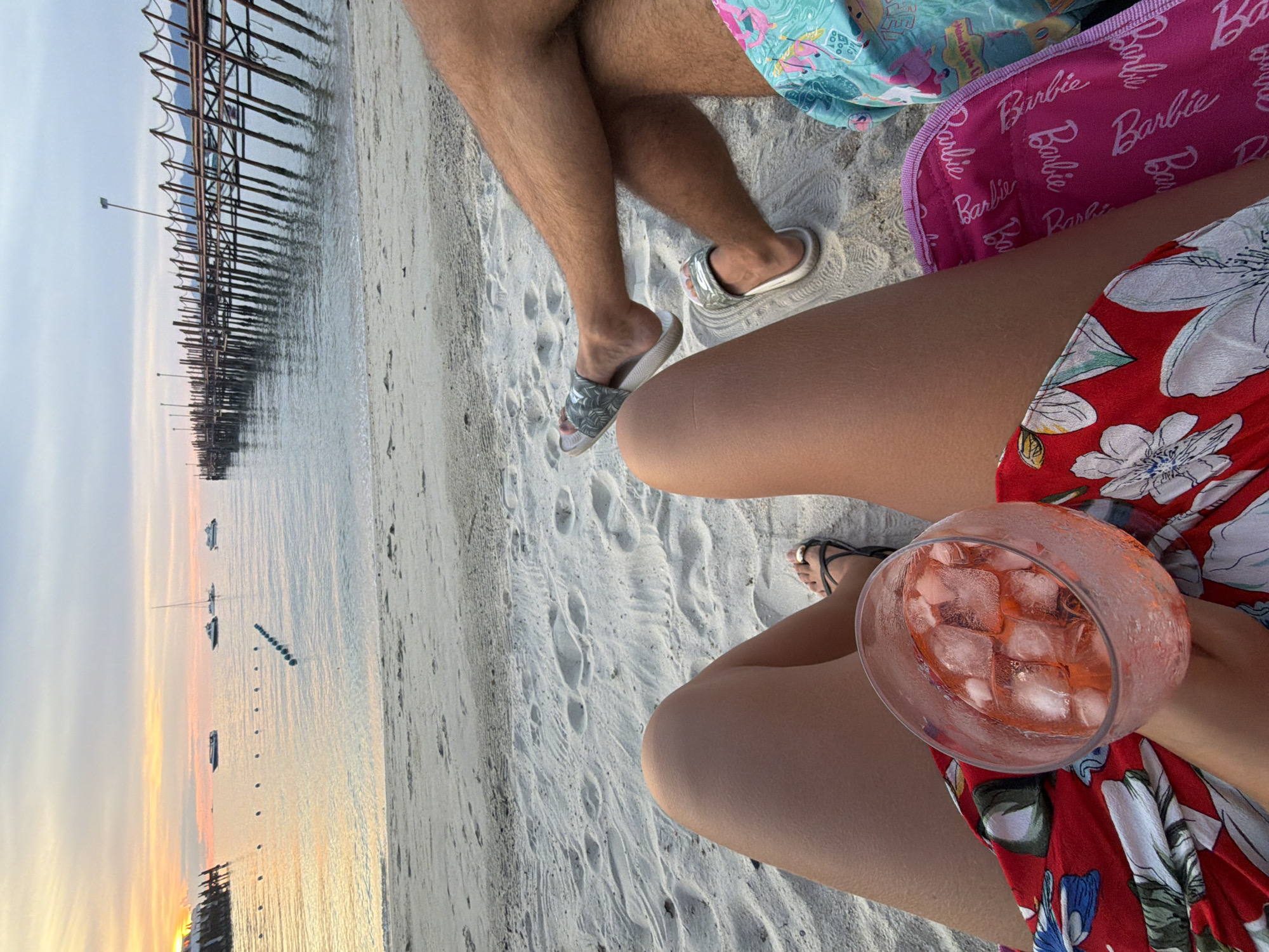 The image depicts a beach scene in Baan Bang Rak, Thailand. In the foreground, two individuals are seated on the sand. One person's legs are visible, holding a glass of a pink beverage with ice. They are wearing a red floral dress and black sandals with a gold ring on their toe. The other person's legs are also visible, crossed at the knees, wearing light-colored slides and patterned swim trunks. The beach sand is light in color and shows footprints. In the background, the calm ocean stretches to the horizon, where a gentle sunset casts warm hues across the sky. Several boats are visible on the water, along with a wooden pier extending into the sea. Buoys mark a lane in the water. The atmosphere suggests a relaxed evening by the coast.