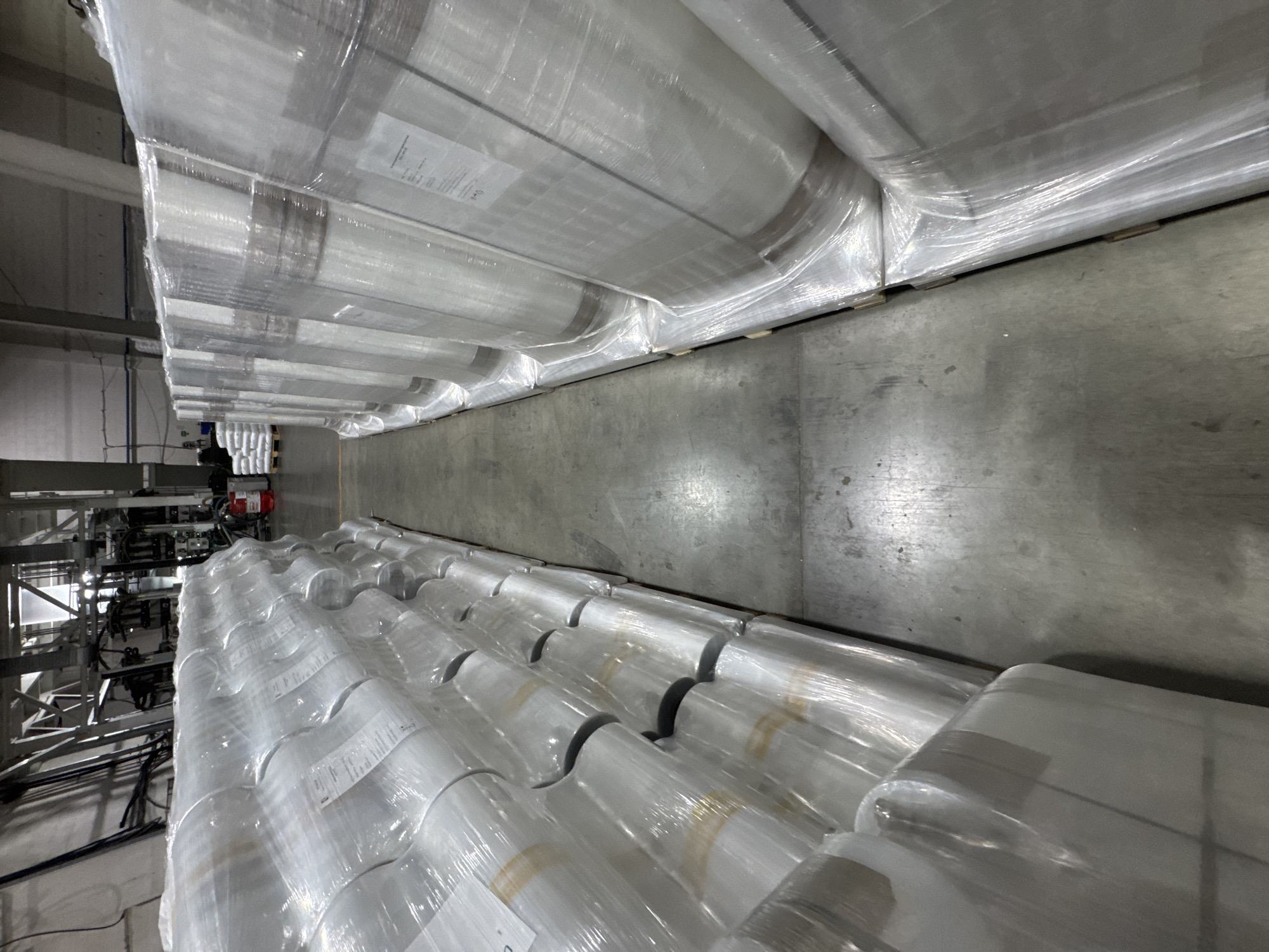 This image depicts an industrial warehouse or factory interior in Perm, Russia, showcasing vast storage and manufacturing operations. The scene is dominated by two long, high rows of stacked materials, forming an aisle that recedes into the distance. Each stack consists of numerous large, white rolls, likely plastic film or similar industrial material, individually wrapped in clear protective plastic. These rolls are arranged on what appear to be pallets or wooden supports.
The floor is a utilitarian gray concrete, showing some signs of wear. In the background on the left side of the aisle, a complex array of industrial machinery is visible, characterized by metal frameworks, wires, and a prominent red motor or component. Faintly, a single person can be seen near this machinery, suggesting an ongoing work activity such as operation or supervision. Labels, which contain indistinct text, are affixed to some of the wrapped rolls, likely indicating product specifications, batch numbers, or other inventory information. The overall impression is that of a functional, organized industrial environment, focused on production or storage of goods. The lighting is consistent with an indoor setting, likely from artificial sources.