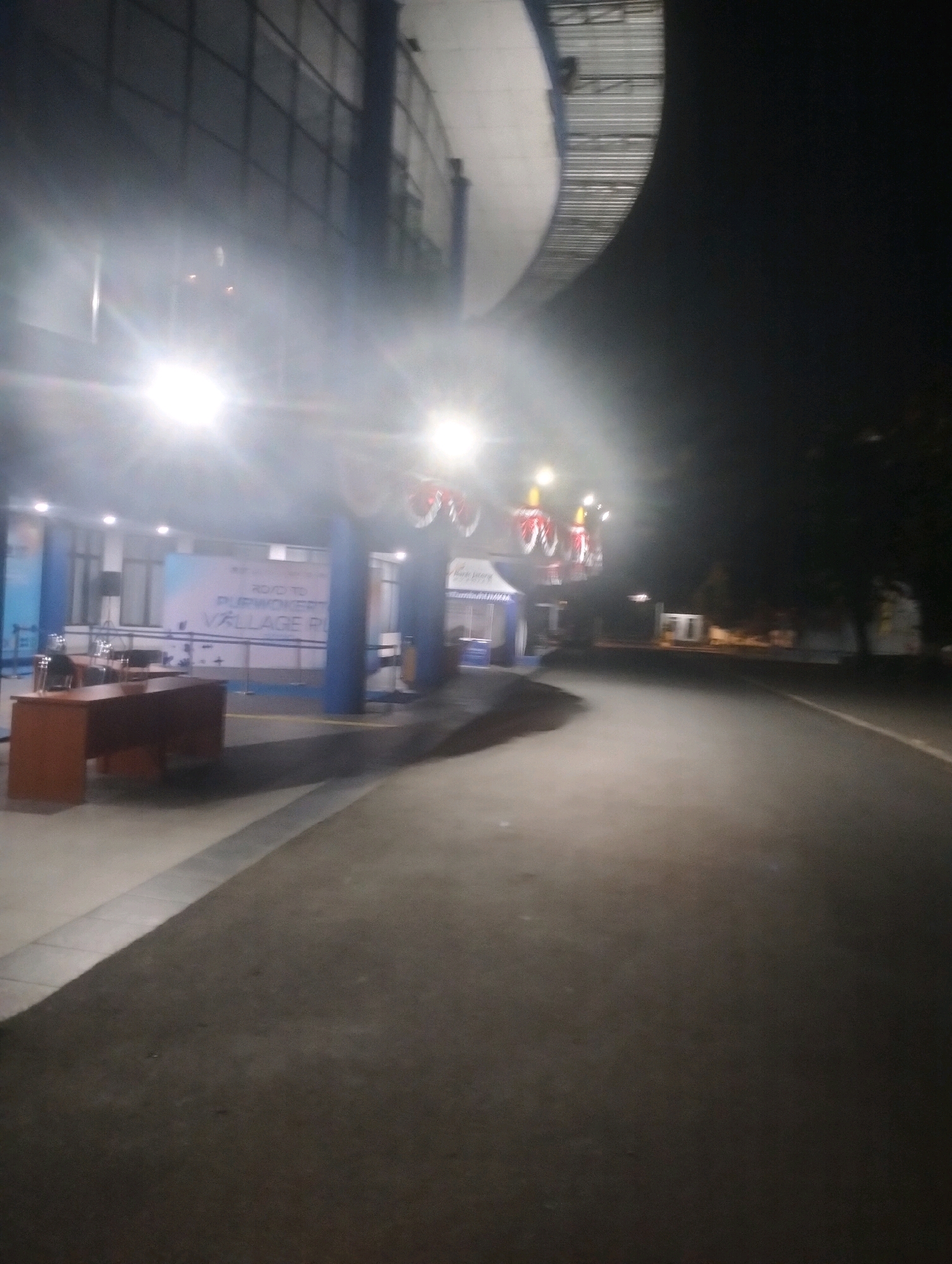 The image depicts a nighttime scene on a paved road next to a building with a curved, metallic roof. The building has a facade with a grid pattern and is illuminated by bright lights. In the foreground, a section of a building's interior is visible, featuring a reception desk, chairs, and a banner with text that appears to read "ROAD TO PURWOKERTO VILLAGE RUN". Red and white decorative elements are strung across the exterior of the building. The road surface is dark asphalt, and the surrounding environment is dark, suggesting it is late at night. The scene is located in Purwokerto, Indonesia.