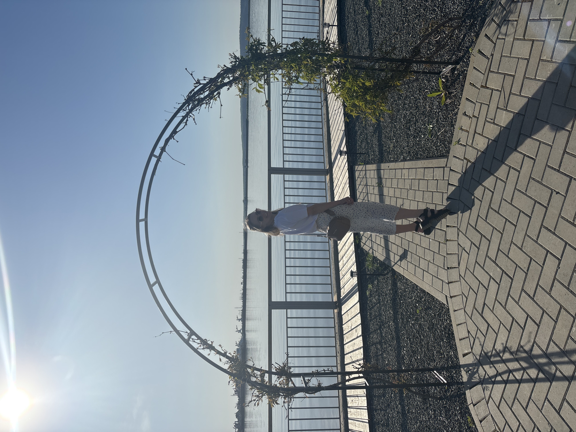 A medium shot captures a woman standing on a brick-paved area, with a body of water and horizon visible in the background. The location is near the city of Chaikovsky, Russia. The woman is wearing a white t-shirt, a patterned skirt, and sandals. She is holding a tan purse in her left hand. A metal archway, partially covered with vines, frames the scene. A fence separates the paved area from a gravel bed. The water body extends to the horizon under a clear sky. The sun shines brightly from the left of the frame.