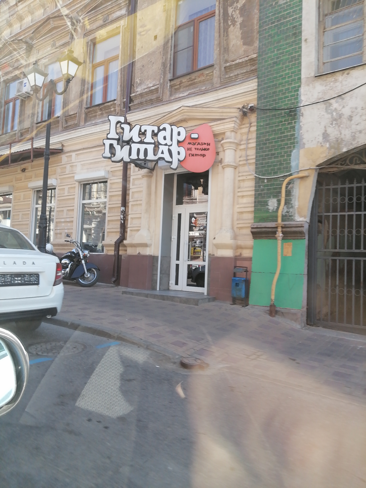 The image captures a street scene in Rostov-on-Don, Russia, focusing on the exterior of a building with a sign reading "ГИТАР" (Guitar). The building features ornate architectural details and large windows. A street lamp with multiple lights is visible. In front of the building, a dark-colored motorcycle is parked. To the left, the rear of a white car is visible, with a license plate that appears to be Russian. The sidewalk is paved with brick-like material. In the background, a green tiled section of the building is present, along with a yellow pipe and a metal gate. The lighting suggests it is daytime, with sunlight casting shadows and reflections. The overall impression is that of a commercial street with a focus on a music-related establishment.