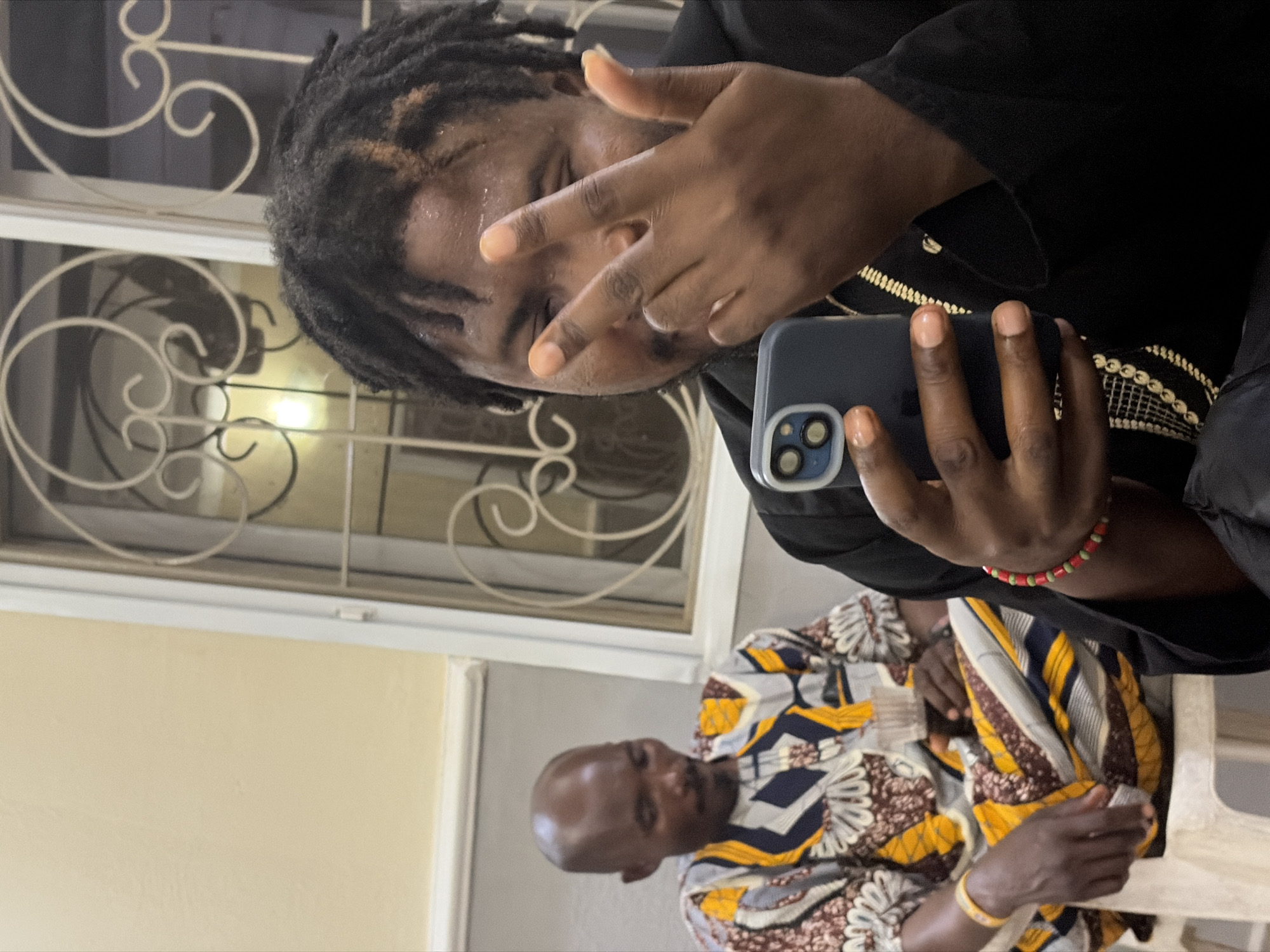 A partially visible man with dreadlocks is holding a smartphone, obscuring his face with a "peace" sign gesture. The phone has a dual-lens camera setup. He is wearing a dark garment with gold trim. Below him, another man with a shaved head is seated, wearing a colorful patterned shirt. The environment includes a white-framed window with decorative ironwork and a plain wall. The location is Jos, Nigeria.