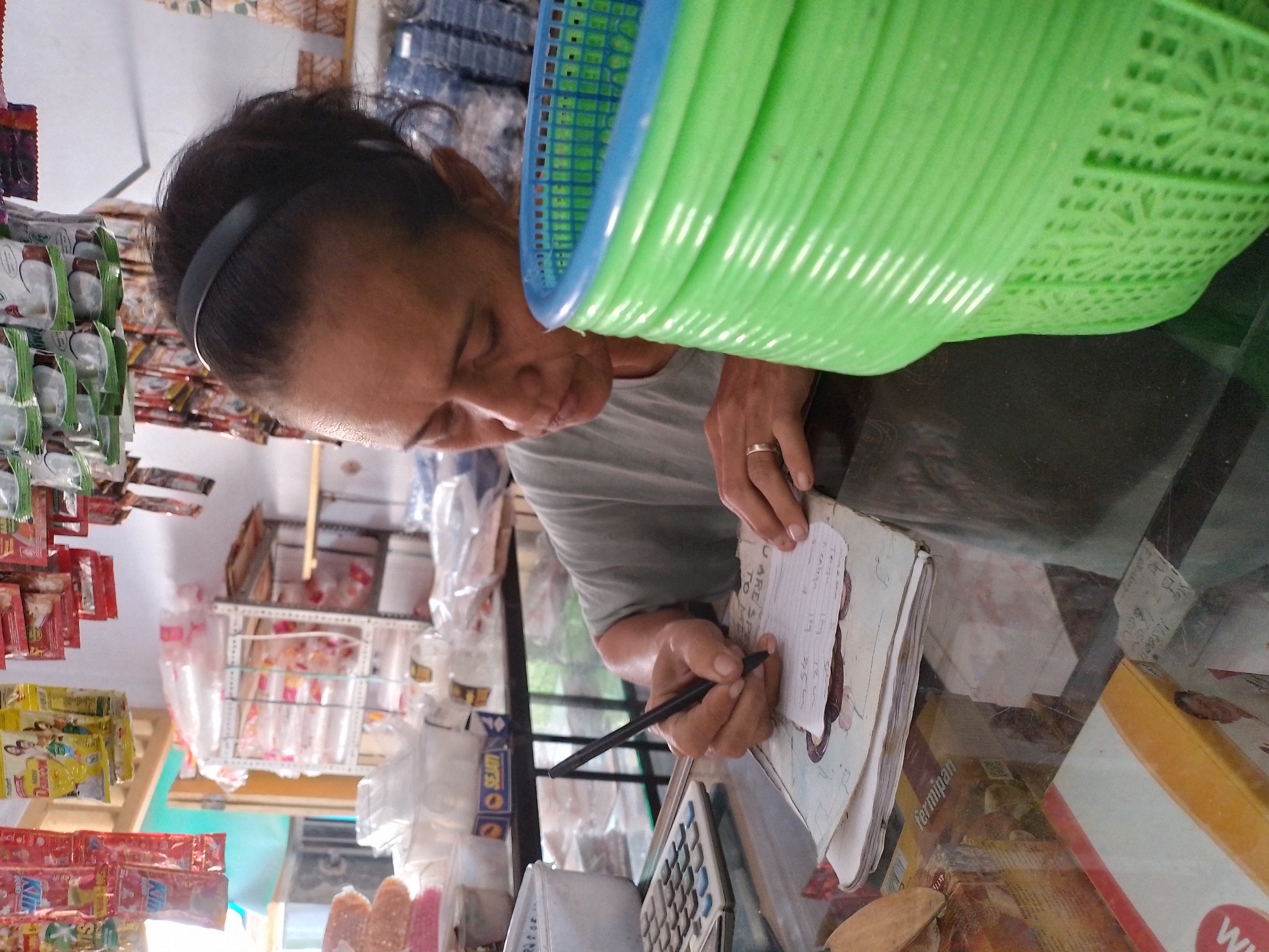 A woman is writing on a piece of paper in what appears to be a small shop in Ngancar, Indonesia. She is wearing a grey t-shirt and has a black headband in her dark hair, pulled back from her face. She is wearing a ring on her left hand, which is holding a black pen and writing on a stack of papers. To her left, a calculator is visible. In the foreground, a bright green plastic basket with a blue rim is stacked. Behind the woman, shelves are stocked with various items, including packaged goods and plastic containers. The overall impression is a scene of daily commerce in a local Indonesian setting.