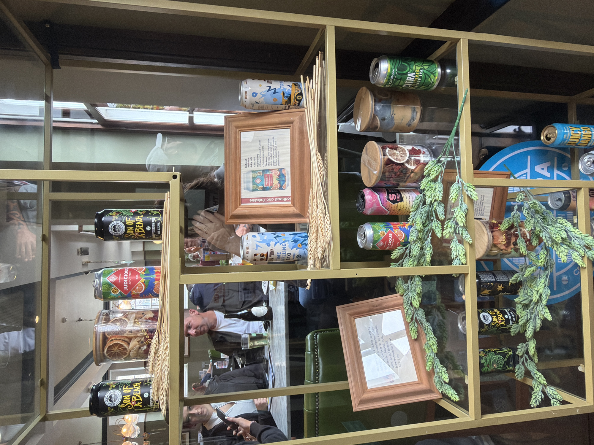 An indoor scene displays a gold-framed shelving unit filled with various items, including canned beverages, jars, and decorative elements. The shelving unit is positioned in what appears to be a public establishment, possibly a bar or restaurant, with people visible in the background.
The top shelf features two cans of "Son of a Beach" beer, a jar filled with dried citrus slices and other items, and a can labeled "JUDECIOUS JUICY PALE." Several stalks of wheat are arranged in front of these items.
The second shelf contains a framed picture with text and an illustration of a beer can, a tall can with a blue and yellow design and "NEW YORK TRAVELLING TAXI" text, and a can with a red and green design. Decorative green hop sprigs are placed in front of the framed picture and the cans.
The third shelf has two small jars with wooden lids, one containing dried fruit slices and the other a beige substance, alongside a can of "OAKHAM CITRA SESSION IPA." More green hop sprigs are displayed here.
The bottom shelf shows several more cans of beer, including "Son of a Beach" and cans with green and black designs. A large blue circular object with white lettering is also visible. Additional green hop sprigs are present.
The overall lighting suggests an indoor environment with ambient light, possibly from windows or overhead fixtures. The establishment appears to be busy, with patrons seated at tables in the background and staff members attending to them. The location is identified as being in Epsom and Ewell, United Kingdom.