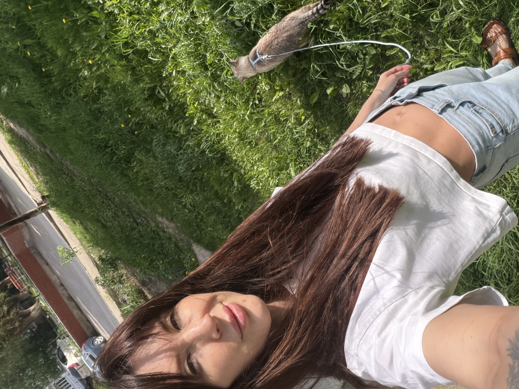 A woman with long brown hair lies on a grassy lawn next to a tabby cat on a leash. The woman is wearing a white t-shirt and light blue jeans, with brown sandals on one foot. The location is Perm, Russia. A sidewalk and buildings are visible in the background.