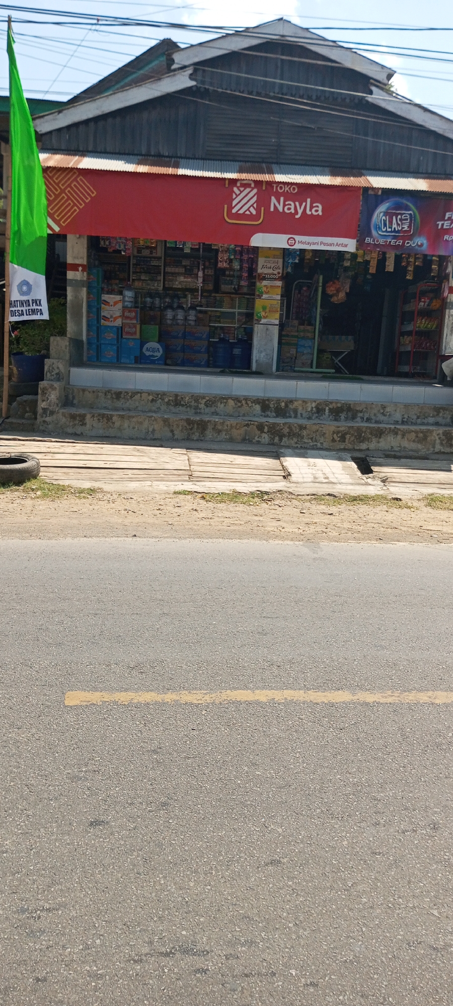 The image shows a small storefront in the city of Lempa, Indonesia, with the store name "TOKO Nayla" visible. The store appears to be a convenience store or warung, selling various goods displayed on shelves behind a counter. There is a green flag on the left side of the image with text that reads "HATINYA PKK DESA LEMPA," which suggests it is related to a community program in Desa Lempa. A tire is visible on the ground to the left of the store entrance. The store is situated on a street with a paved road and a yellow line marking the lane. The building has a corrugated metal roof and is accessed by a set of concrete stairs. The sky is visible in the background, and it appears to be a clear day.