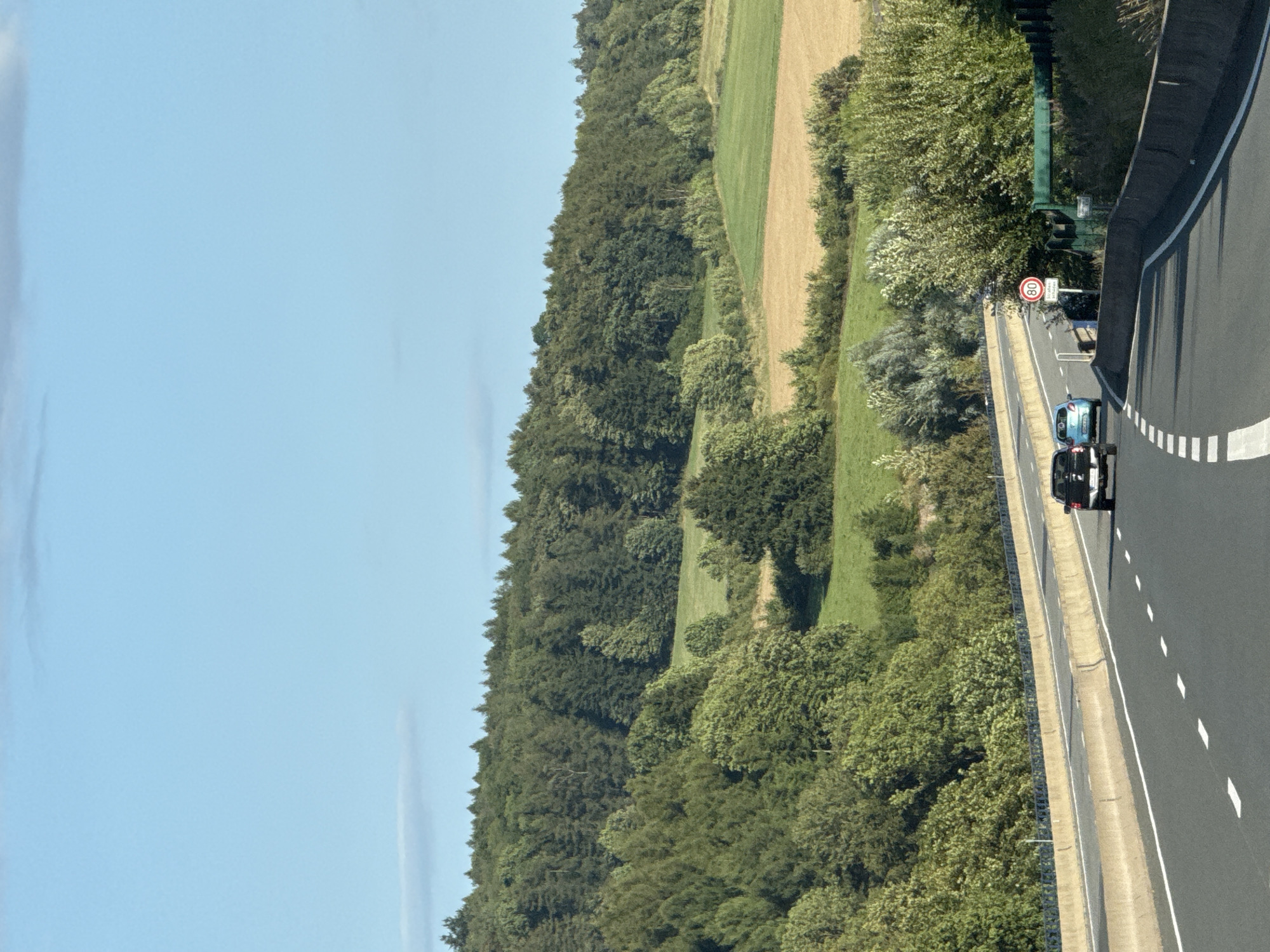 This is a photograph of a highway with a rural landscape in the background. The highway has concrete barriers on either side and is divided into lanes by white dashed lines. There are two cars on the highway: a black pickup truck and a small blue car. Both cars are moving away from the camera. In the background, there is a dense forest with a clear blue sky above. There are also some green fields and a few trees visible in the distance. The scene is brightly lit, suggesting it is daytime.
This image is not located in the city of Bad Lauterberg im Harz, Germany, as the image depicts a rural highway with no recognizable landmarks or signage that would indicate this specific location.
