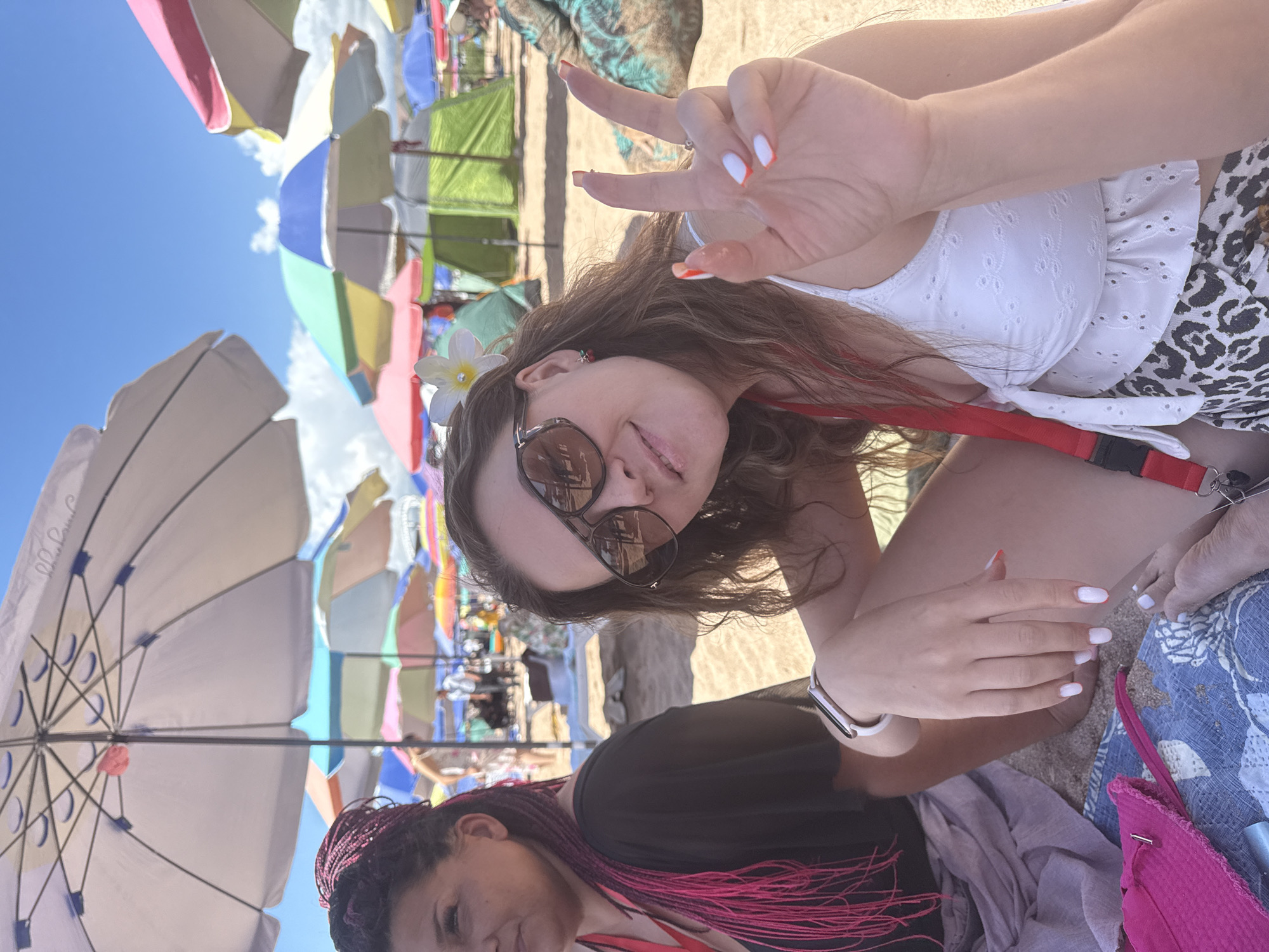 The image depicts two women on a sandy beach, with numerous colorful beach umbrellas in the background. The woman in the foreground, with long brown hair, is wearing sunglasses, a white top, and a leopard print skirt. She is making a peace sign with her right hand, which has white nail polish with orange tips. A white flower is tucked behind her ear. The woman on the left, with dark hair styled in pink braids, is wearing a black top. Both women are seated on the sand, with a patterned mat and a pink bag visible in the lower left corner. The sky is blue with some clouds. The location is identified as Bosteri, Kyrgyzstan.