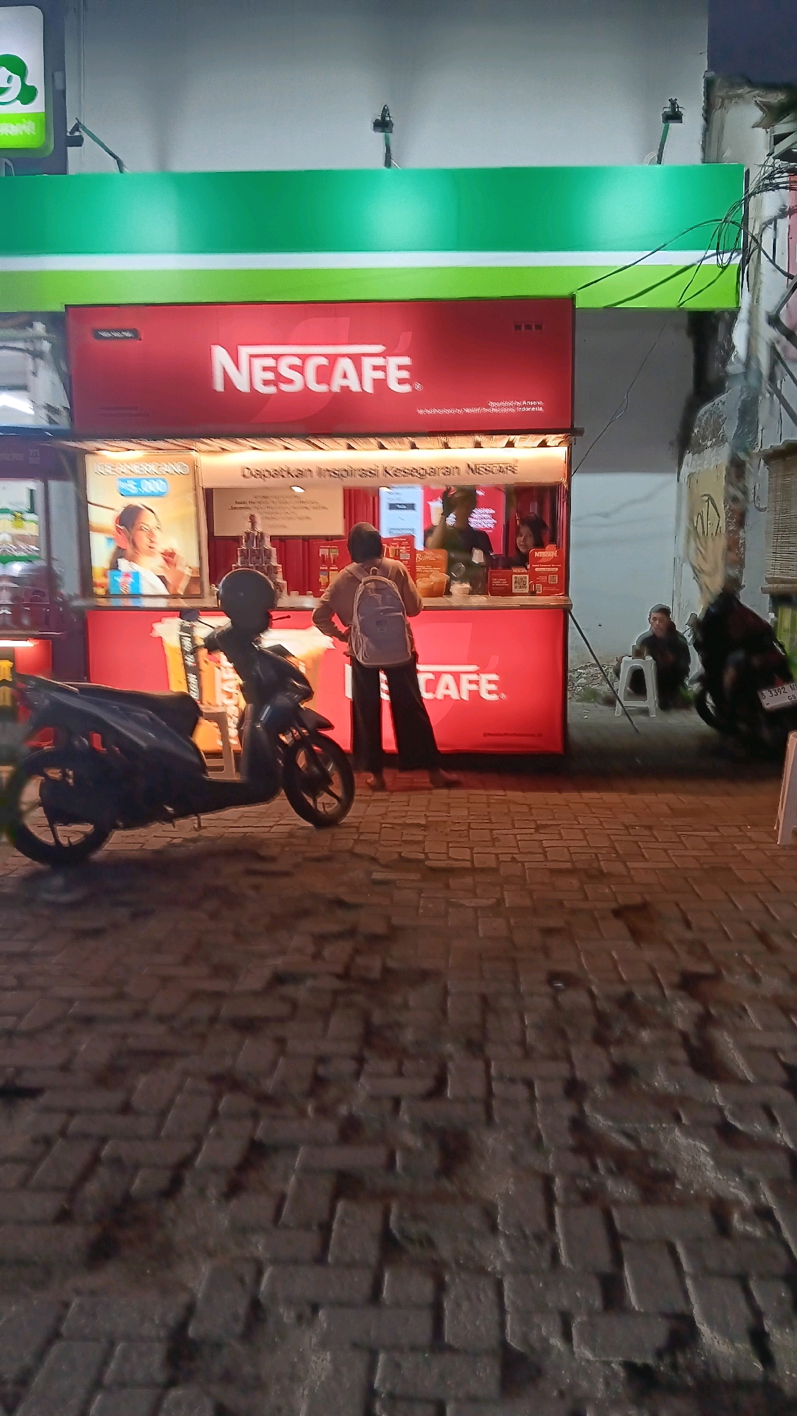 An outdoor street-level shot captures a temporary Nescafé coffee stand at night, located on Jalan Boulevard Bintaro Utama, South Tangerang, Indonesia. The stand features a bright red counter with the Nescafé logo prominently displayed. Behind the counter, two employees are visible. In front of the stand, a customer with a backpack, dressed in dark clothing and white shoes, is interacting with the staff. A dark-colored scooter is parked to the left of the stand. To the right, two individuals are seated on low stools, with a motorcycle parked nearby. The ground is paved with dark brick. A portion of a green awning is visible overhead.