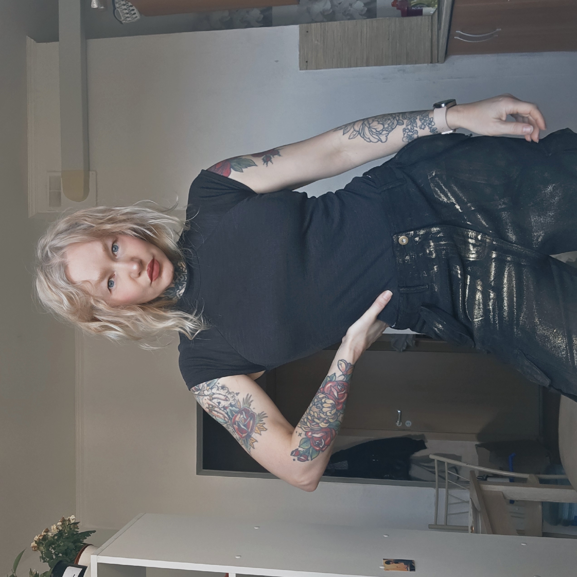 A woman with fair skin, blonde wavy hair, and multiple tattoos on her arms poses for a photograph in an indoor setting. She has blue eyes and red lipstick, and her attire consists of a black turtleneck shirt and dark pants with a gold shimmer. Her pose involves her left hand resting on her hip.
The setting appears to be a room with light-colored walls. In the background, there is a white storage unit, and near it is a potted plant with wilting white flowers. A dark-framed doorway is visible, beyond which is a partially visible wooden staircase with white railings, and what appears to be a dark coat or garment. No evidence suggests that the location is in the city of Saint Petersburg, Russia.