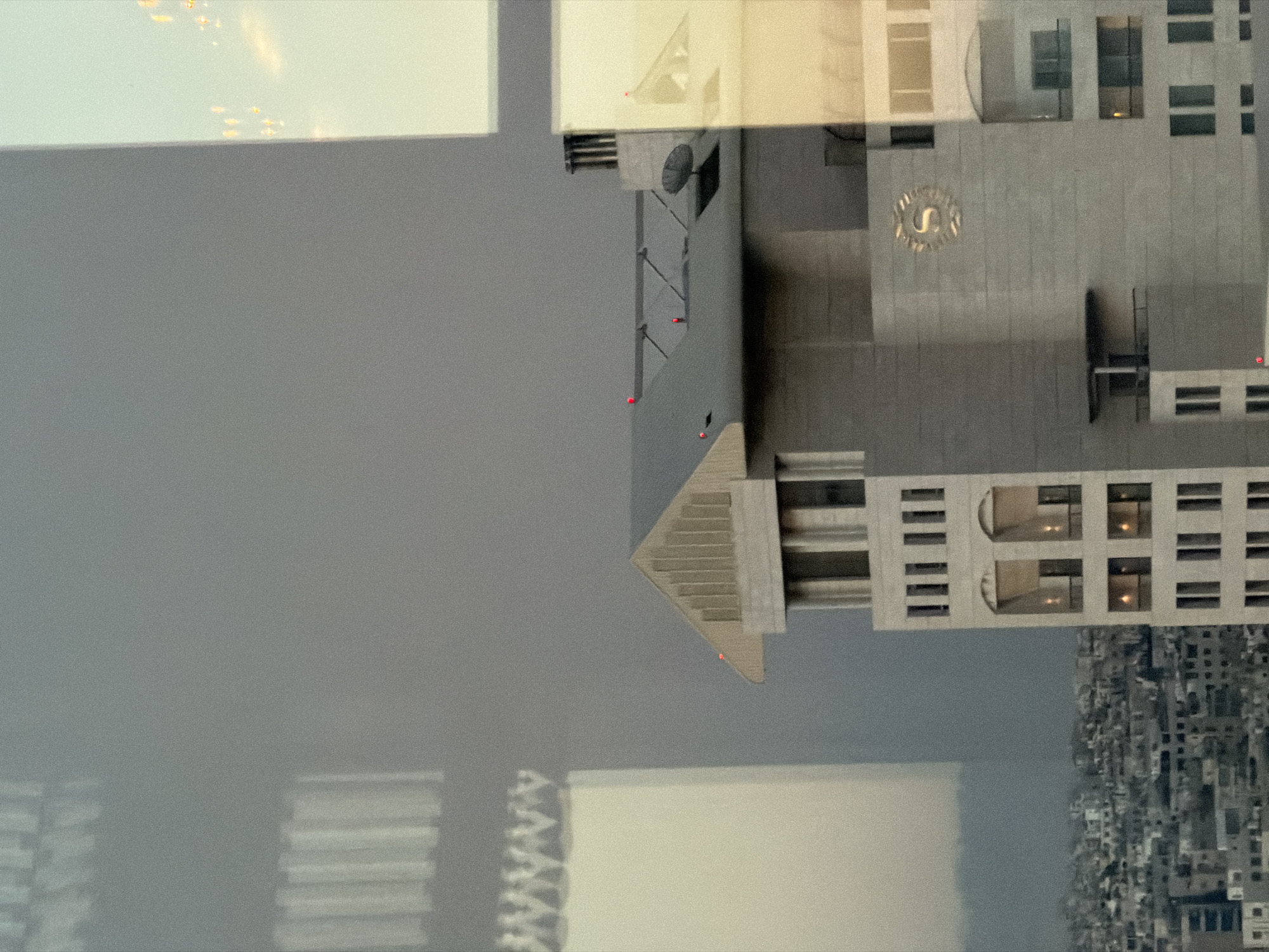 This is a daytime, medium-angle, eye-level outdoor shot of a modern building in Amman, Jordan, viewed through a window with reflections. The primary subject is a light-colored, multi-story building with a prominent "S" logo on its facade, which appears to be the Sheraton Hotel. The building has several floors with distinct window arrangements, including arched openings and rectangular windows. Red lights are visible on the rooftops of this building and another adjacent structure, indicating potential navigational lights or markers.
To the left and background, a densely populated urban landscape is visible, characterized by numerous smaller buildings and structures. A large, somewhat indistinct building or structure with vertical lines is reflected or appears in the foreground on the left, possibly another building or a distorted reflection from the window. The sky is overcast and gray, suggesting either a cloudy day or haze, with some subtle warm light in the upper right quadrant, hinting at the sun's position. The overall impression is one of an urban environment, likely viewed from an elevated position such as another building or hotel room.