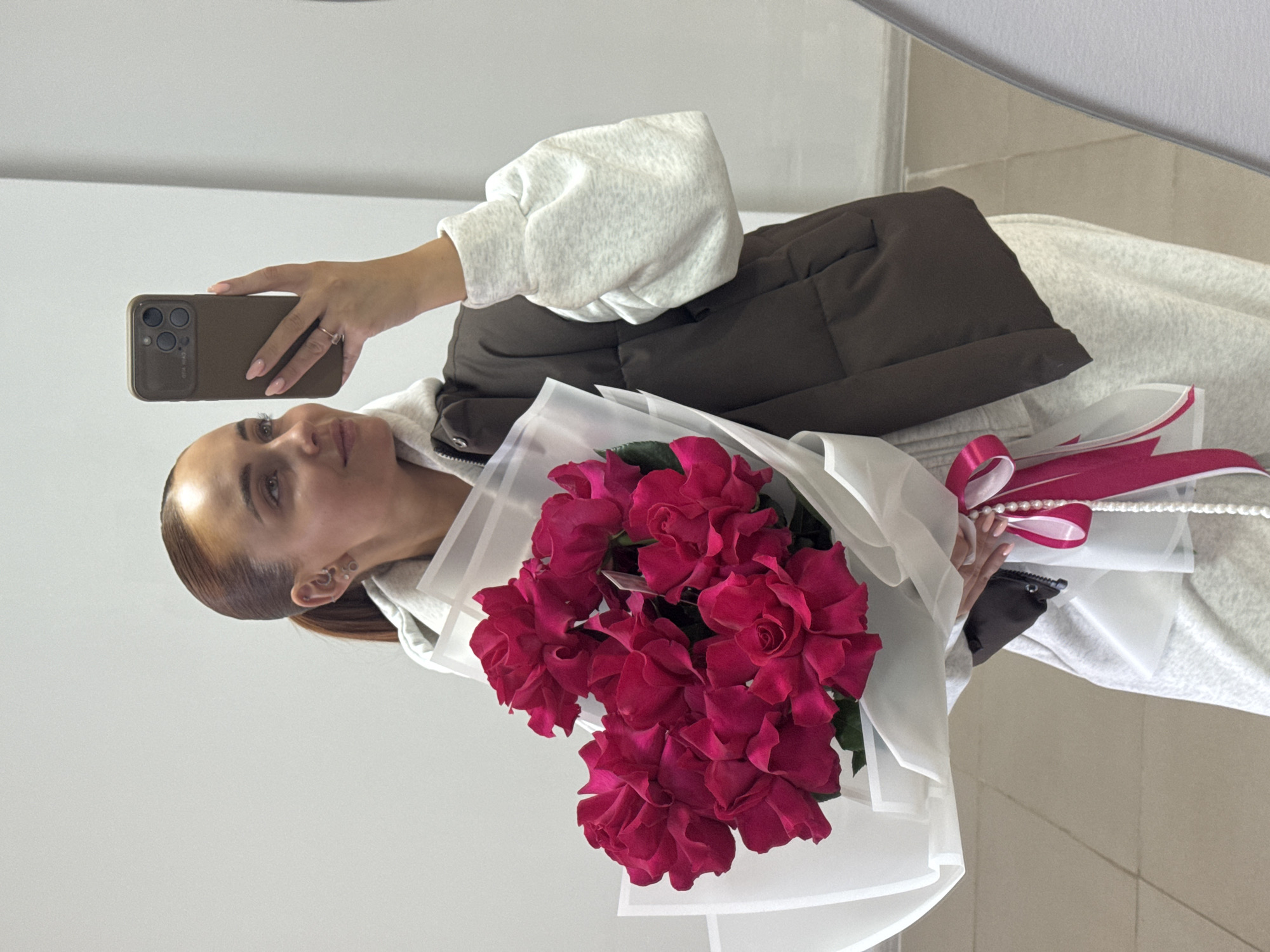 A woman is holding a bouquet of red flowers and taking a picture of herself with a smartphone. She is wearing a white hoodie, a brown vest, and light-colored pants. The bouquet is wrapped in white paper and tied with a pink ribbon and a string of pearls. The background appears to be an interior with plain white walls. The city of Chaikovsky, Russia is not identifiable.