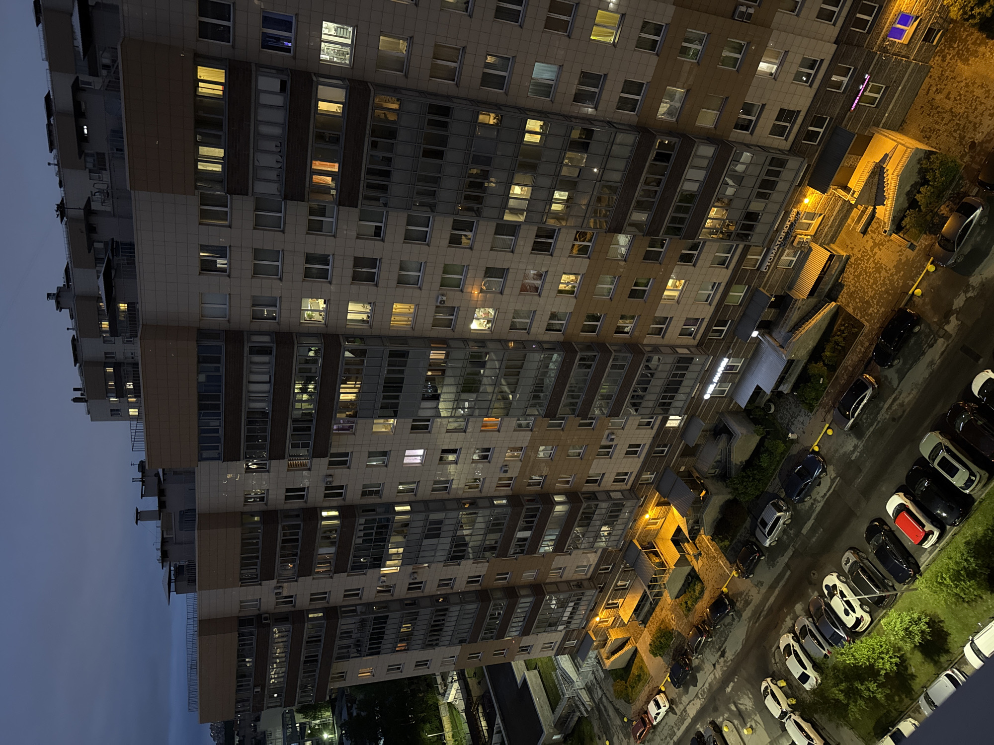 The image shows a tall residential building in Unknown, Russia, at dusk or night. The building has many windows, some of which are illuminated, suggesting that residents are home. Below the building, there is a parking lot filled with cars of various makes and models. Adjacent to the parking lot, there is a small patch of greenery. The sky is dark, indicating that it is either late in the evening or early morning.