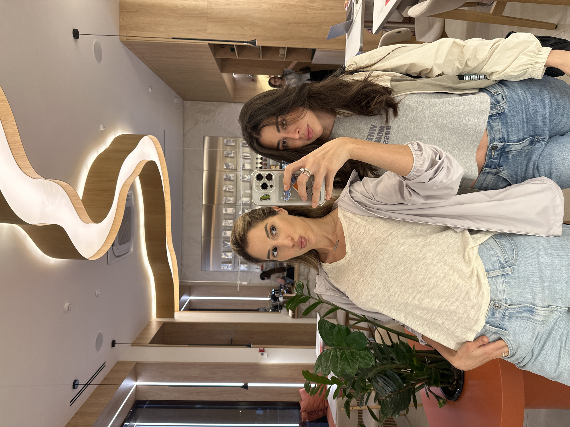 Two young women are taking a mirror selfie in what appears to be a modern cafe in Vladikavkaz, Russia. The woman on the left has long blonde hair and is wearing a light-colored t-shirt, a light purple jacket, and light blue jeans. She is puckering her lips and looking at the camera. The woman on the right has long dark hair and is wearing a grey t-shirt with text, a cream-colored jacket, and blue jeans. She is looking directly at the camera with a neutral expression. The cafe has wooden accents on the walls and a distinctive wavy ceiling light fixture with integrated lighting. There are tables and chairs visible in the background, along with a plant in a pot on an orange table in the foreground. The overall environment suggests a contemporary and stylish establishment.
