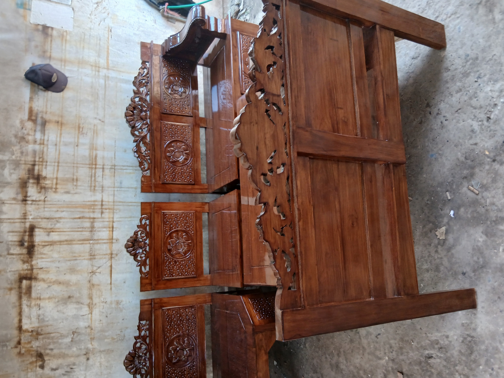 In Tenggarang, Indonesia, a row of ornate wooden chairs and benches with intricate carvings are displayed against a weathered, textured wall. The furniture, crafted from dark, polished wood, features detailed floral motifs and elaborate scrollwork on the backrests and armrests. The chairs appear to be part of a set, with three visible in the foreground and what looks like an additional armrest extending from the right side of the frame. In the background, a brown baseball cap hangs on the wall. The floor is concrete, with scattered debris suggesting an outdoor or workshop setting. The lighting is somewhat dim, highlighting the rich tones of the wood and the craftsmanship of the pieces.