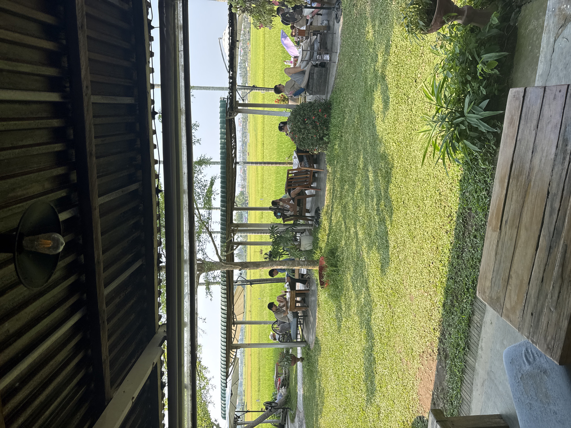 The image shows an outdoor seating area at a venue. Several groups of people are sitting at tables enjoying the view of a rice paddy. The seating area is shaded by a series of wooden structures with awnings. The sky is clear and the sun is shining. In the foreground is a partial view of a wooden table. No evidence confirms the location in Đà Nẵng, Vietnam.